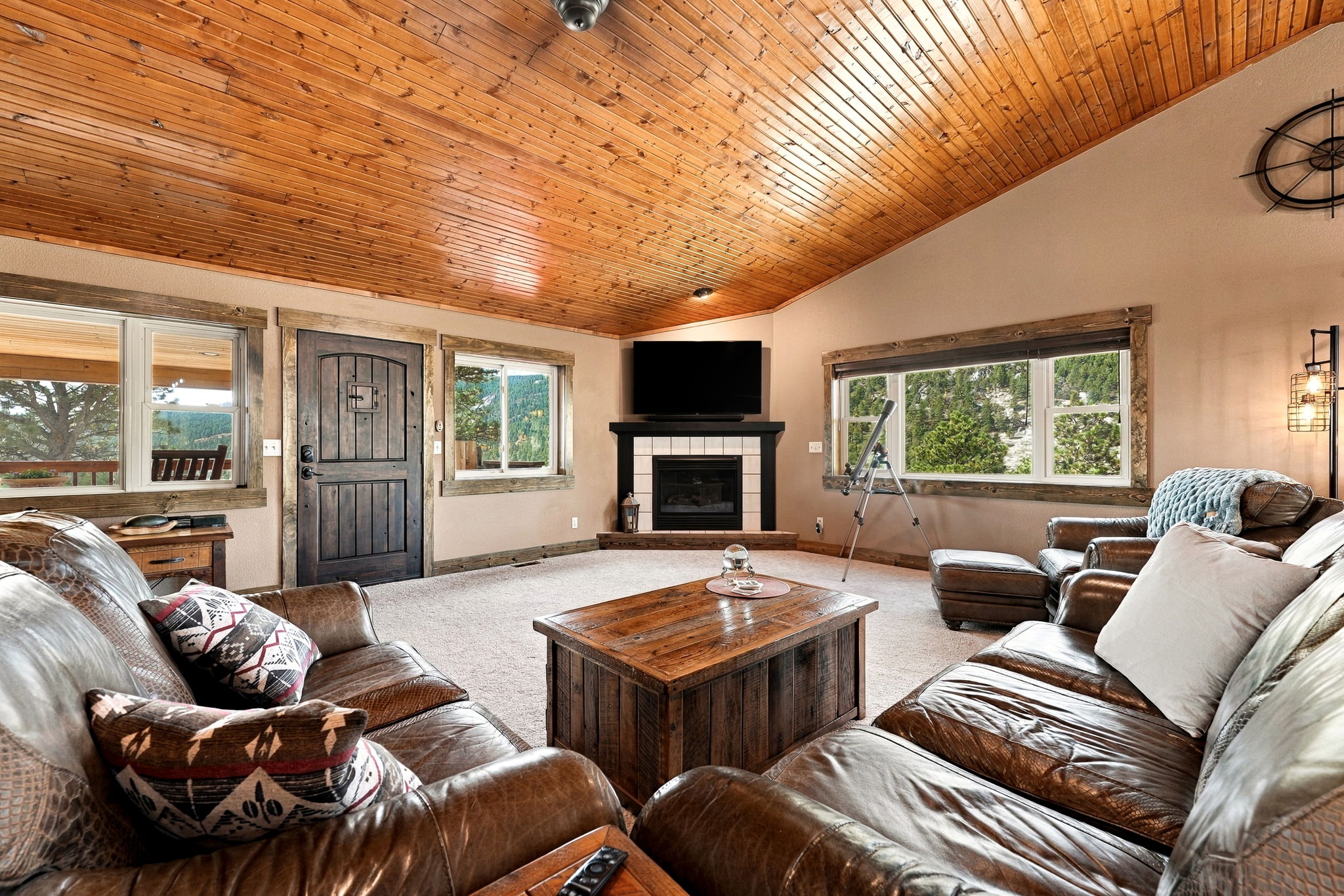 This rustic living room features a vaulted wood-paneled ceiling and a cozy corner fireplace with a dark mantel and tiled surround. The space is furnished with comfortable leather seating, a sturdy wooden coffee table, and large windows that frame scenic mountain views. A telescope positioned near the window adds a unique touch to this inviting, cabin-style retreat.