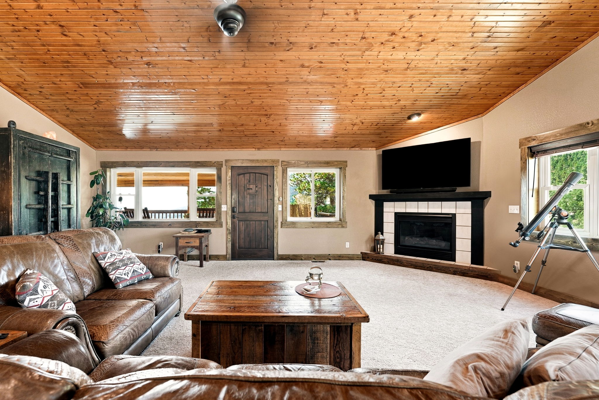This rustic-style living room features a vaulted wood-paneled ceiling and a cozy seating area centered around a fireplace with a black mantel and white tile surround. A large leather sofa and a substantial wooden coffee table anchor the space, while a telescope positioned near a window adds a unique touch. The room is bathed in natural light from multiple windows, creating a warm and inviting atmosphere.