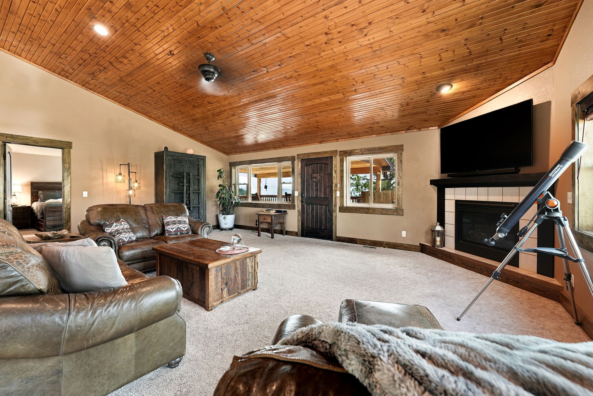 This cozy, rustic-style living room features a vaulted wood-paneled ceiling and neutral carpeting throughout. The space is anchored by a fireplace with a dark mantel and a television mounted above, while a leather sofa set and a wooden coffee table provide comfortable seating. A telescope stands near the fireplace, and a doorway leads to an adjacent bedroom, creating an inviting and warm atmosphere.