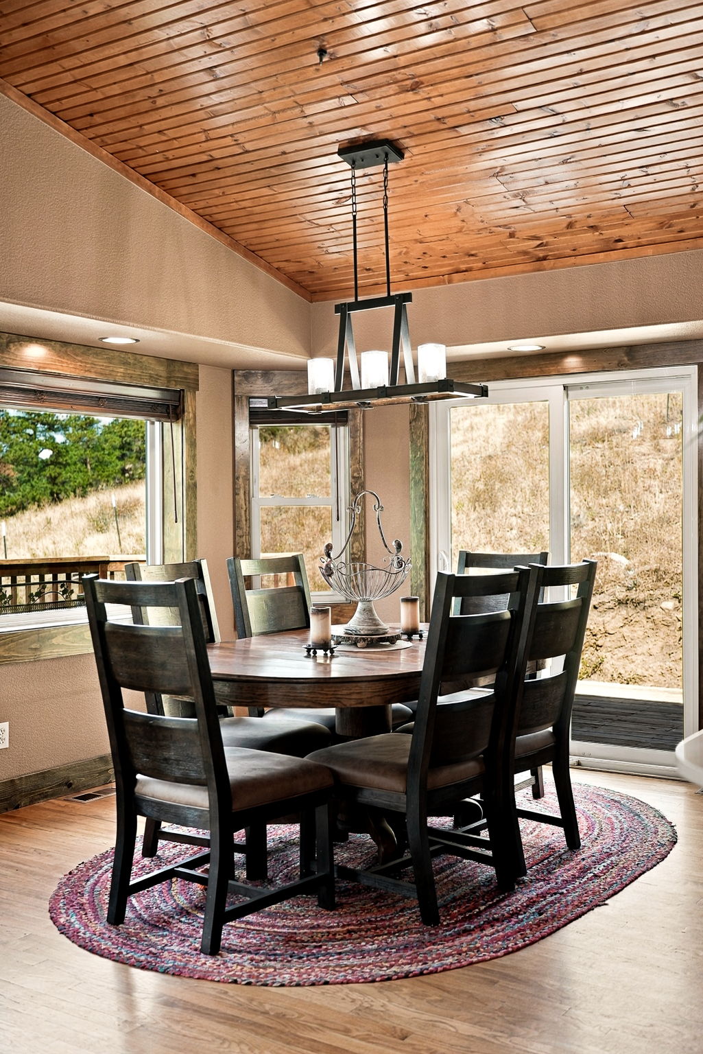 This rustic-style dining room features a dark wood table surrounded by matching chairs, set atop a colorful, oval-shaped braided rug. The space is defined by a warm wood-paneled ceiling and large windows that provide natural light and views of the surrounding landscape. A prominent industrial-style chandelier hangs above the table, creating a cozy and inviting atmosphere for meals.