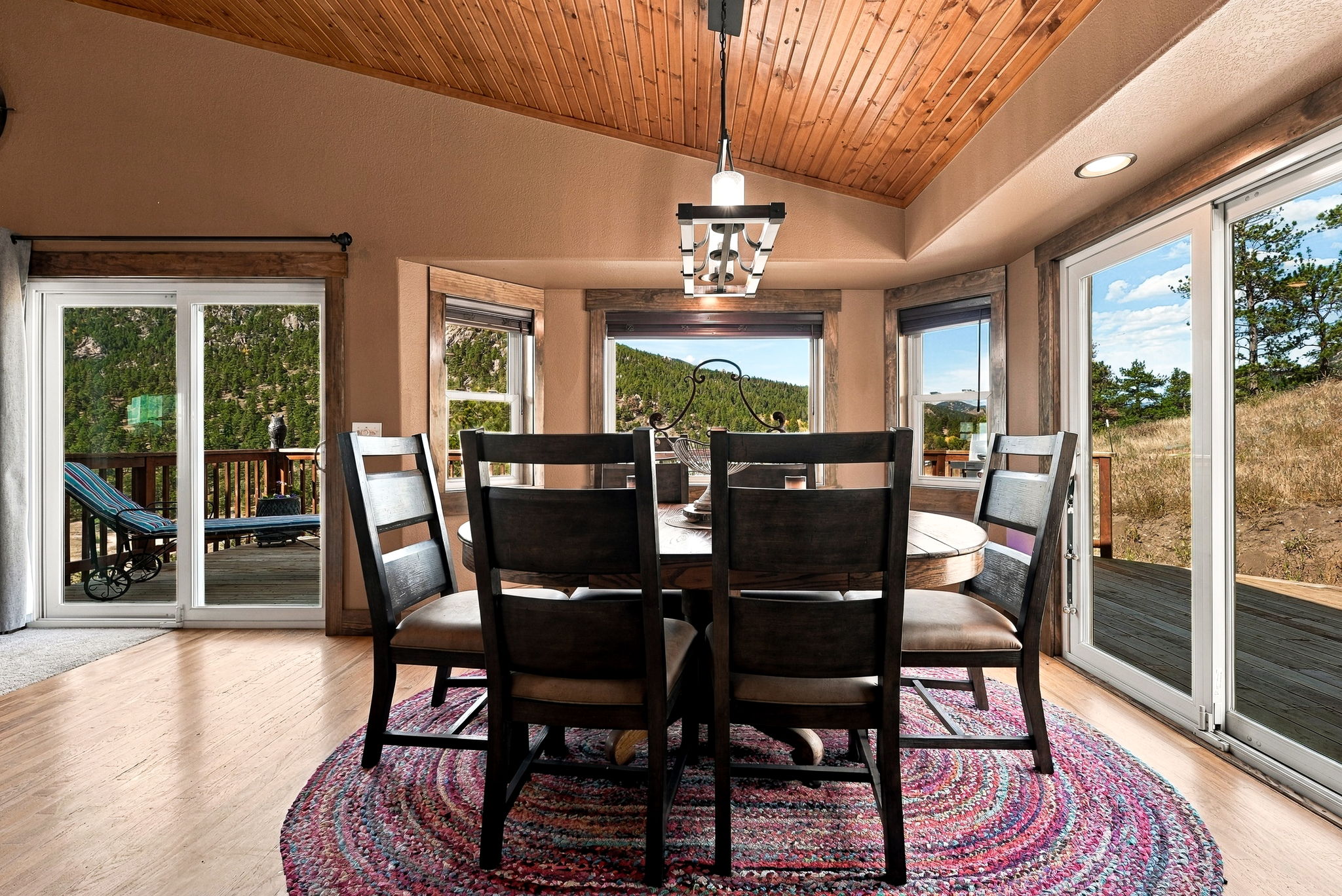 This dining room features a round wooden table surrounded by four dark-toned chairs, all set atop a vibrant, circular patterned rug. The space is defined by a vaulted wood-paneled ceiling and large sliding glass doors on either side that provide expansive views of the surrounding mountain landscape. A modern, multi-light chandelier hangs centrally above the table, creating a warm and inviting atmosphere.