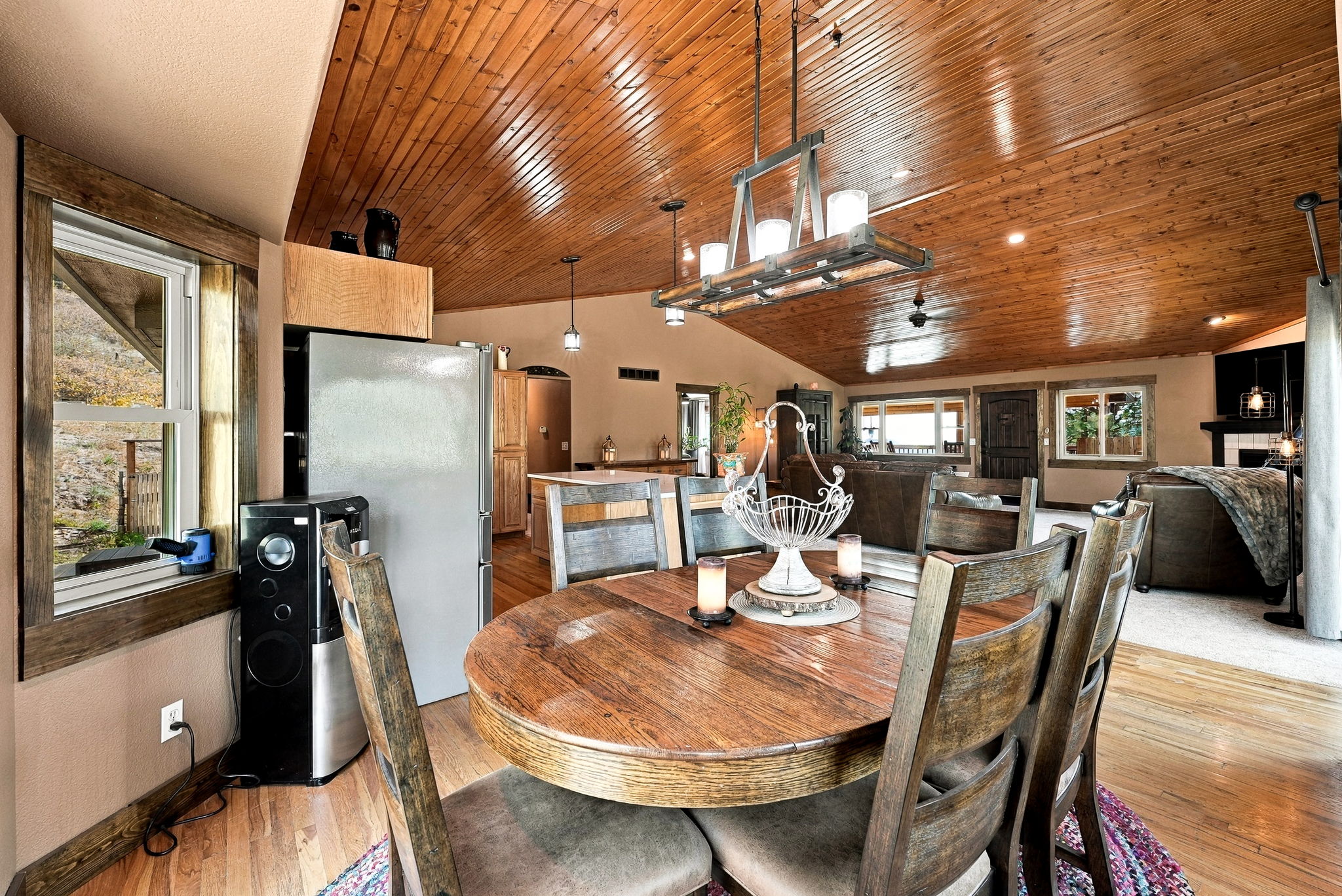 This dining area features a rustic round wooden table surrounded by matching chairs, positioned beneath a distinctive industrial-style chandelier with exposed bulbs. The room showcases a warm aesthetic with wood-paneled ceilings and hardwood flooring, seamlessly transitioning into an open-concept living space. A large window to the left provides natural light, while the overall atmosphere feels cozy and inviting with a blend of traditional and cabin-inspired design elements.