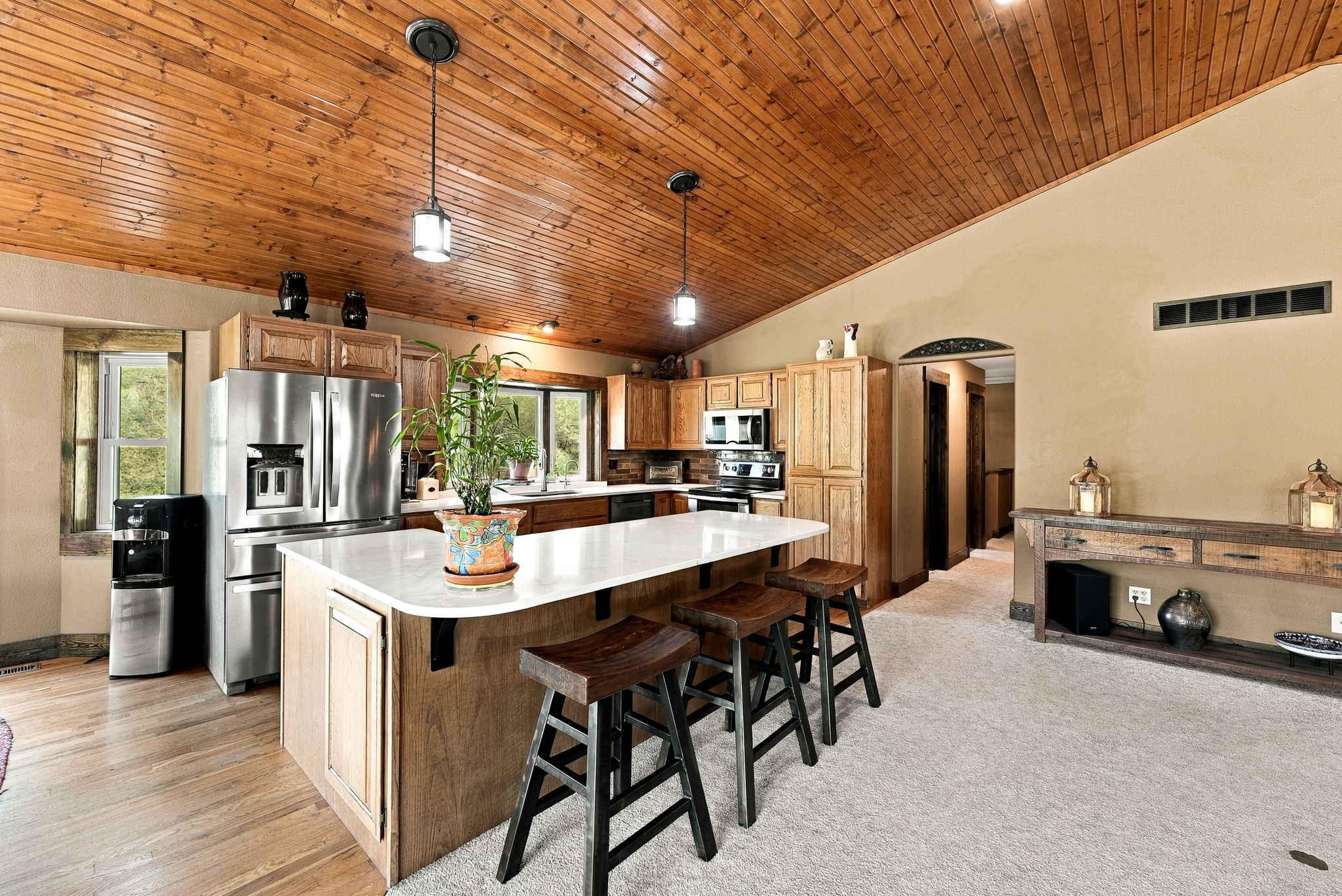 This kitchen features a prominent island with a light-colored countertop and three dark wood stools, set against a backdrop of wood cabinetry and stainless steel appliances. The room is defined by a vaulted wood-paneled ceiling and a mix of wood and carpeted flooring, creating a warm, rustic atmosphere. The perspective captures the open-concept layout, showing the kitchen flowing into an adjacent hallway and a console table area.
