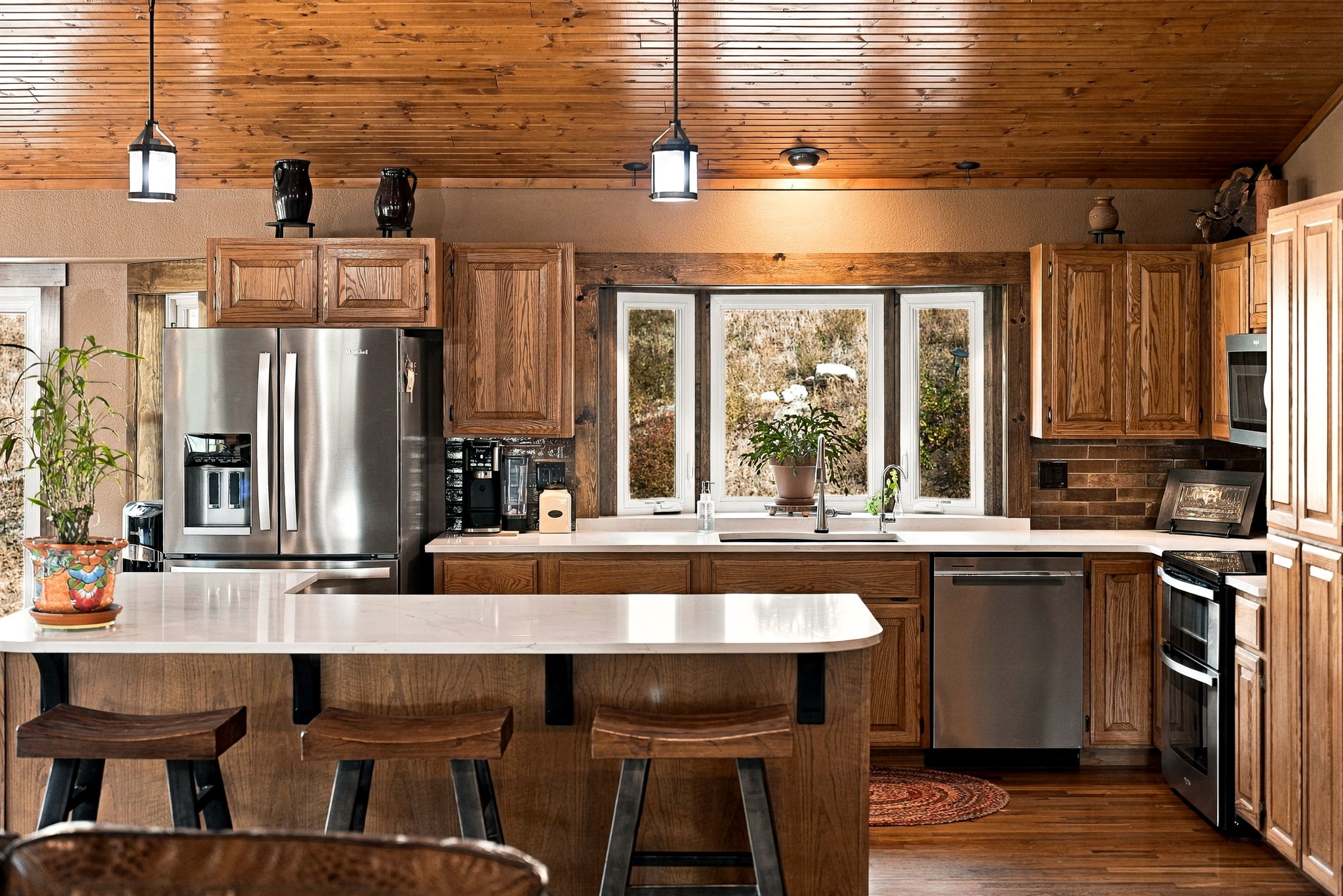 This rustic-inspired kitchen features warm wood cabinetry and a wood-paneled ceiling, creating a cozy, cabin-like atmosphere. A large island with a light-colored countertop and bar seating serves as a central focal point, complemented by stainless steel appliances and a triple-pane window above the sink. The space is well-lit by pendant lights and natural light, offering a functional and inviting layout for cooking and entertaining.