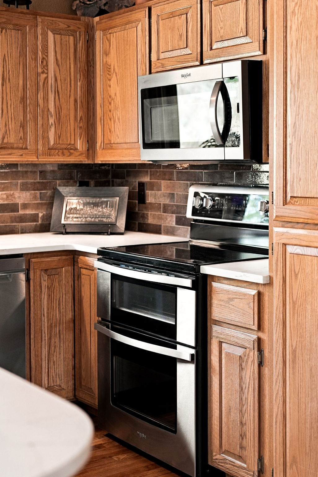 This kitchen features warm-toned wood cabinetry paired with a dark, textured tile backsplash and light-colored countertops. The space is equipped with stainless steel appliances, including a double-oven range and an over-the-range microwave. The perspective is a close-up, eye-level shot that highlights the functional layout and the contrast between the wood finishes and the modern appliances.