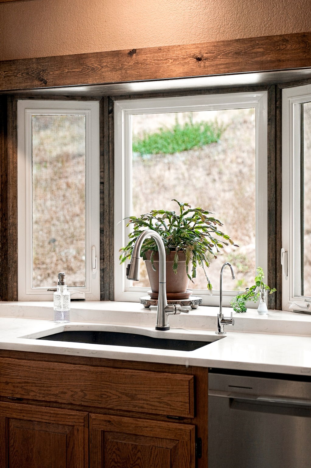 This kitchen features a bright, window-lined sink area with a rustic wood-trimmed bay window overlooking an outdoor landscape. A sleek, modern faucet and a secondary filtered water tap are mounted on a light-colored stone countertop above a dark, undermount sink. The warm wood cabinetry below and the natural light create a cozy, inviting atmosphere perfect for a home-focused real estate presentation.