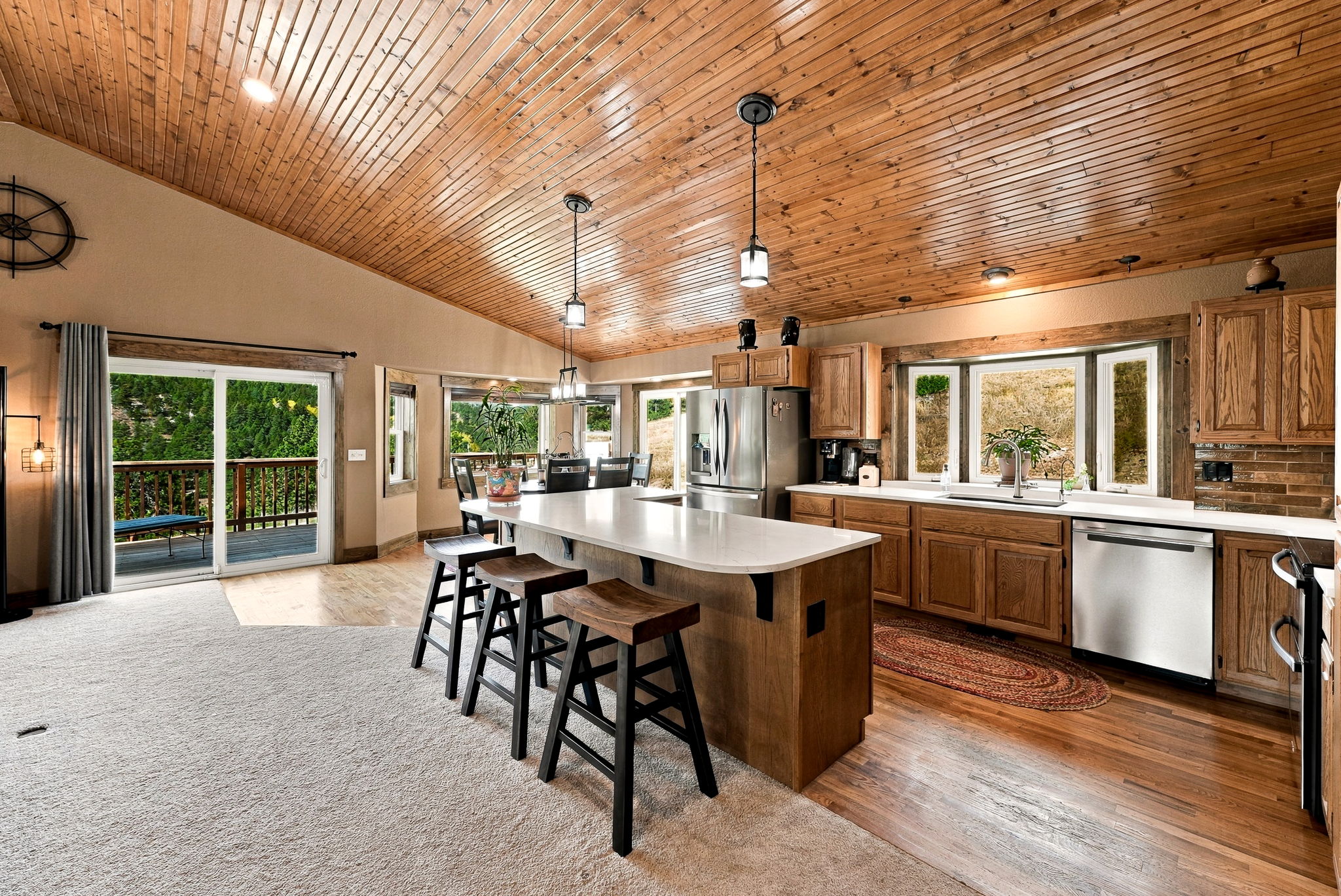 This open-concept kitchen features a large central island with a light-colored countertop and three bar stools, set against a backdrop of warm wood cabinetry and a vaulted wood-paneled ceiling. The space flows seamlessly into a carpeted living area and features large sliding glass doors that provide a view of the outdoor deck. Natural light fills the room through multiple windows, highlighting the blend of rustic wood textures and modern stainless steel appliances.