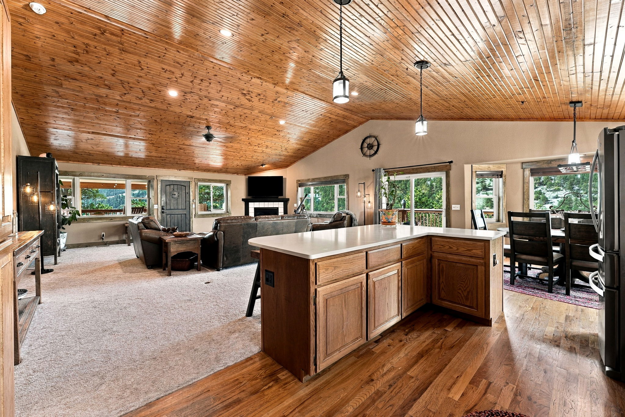 This open-concept living space features a prominent kitchen island with a light-colored countertop, serving as a central hub between the kitchen and the living area. The room is characterized by a vaulted ceiling finished with warm-toned wood planks, complemented by a mix of carpeted and hardwood flooring. Large windows and sliding glass doors provide ample natural light and views of the surrounding greenery, creating a spacious and inviting atmosphere.