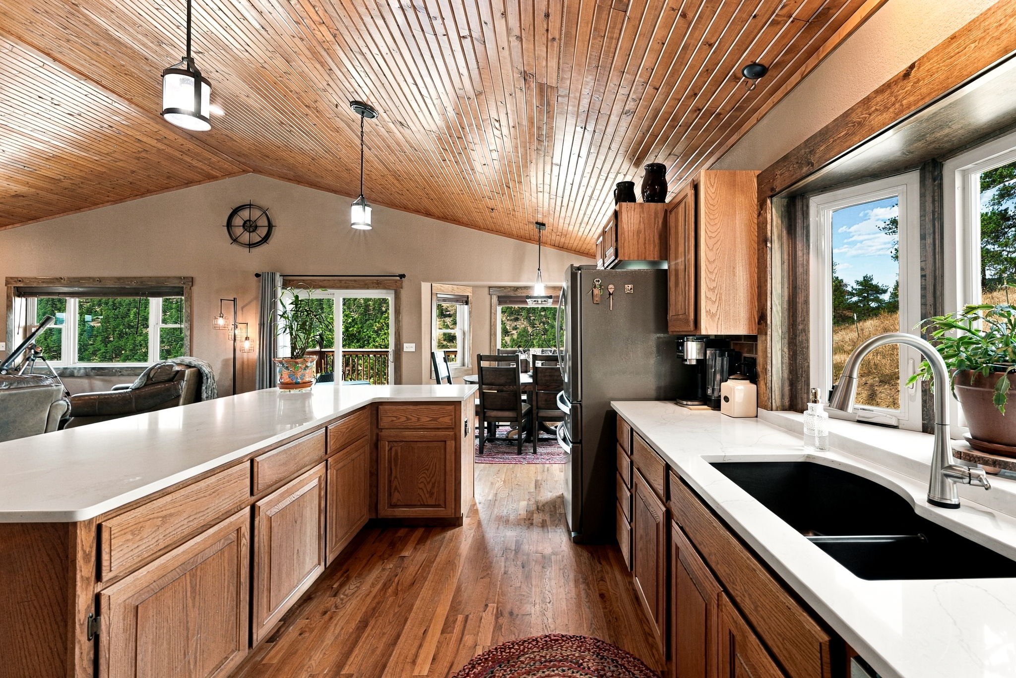 This warm and inviting kitchen features rich wood cabinetry and light-toned countertops, creating a classic rustic aesthetic. The space is anchored by a large island and a deep sink positioned beneath a window that provides a scenic view of the outdoors. The vaulted wood-paneled ceiling adds architectural interest, while pendant lighting illuminates the workspace, leading the eye toward the adjacent dining area.