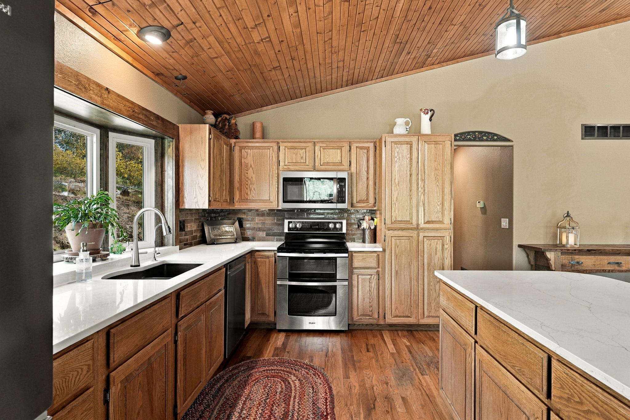 This kitchen features warm-toned wood cabinetry paired with light-colored countertops and a dark tiled backsplash. The space is highlighted by a vaulted wood-paneled ceiling and a large window above the sink that brings in natural light. A central island provides additional workspace, creating a functional and inviting atmosphere with a rustic, cozy aesthetic.