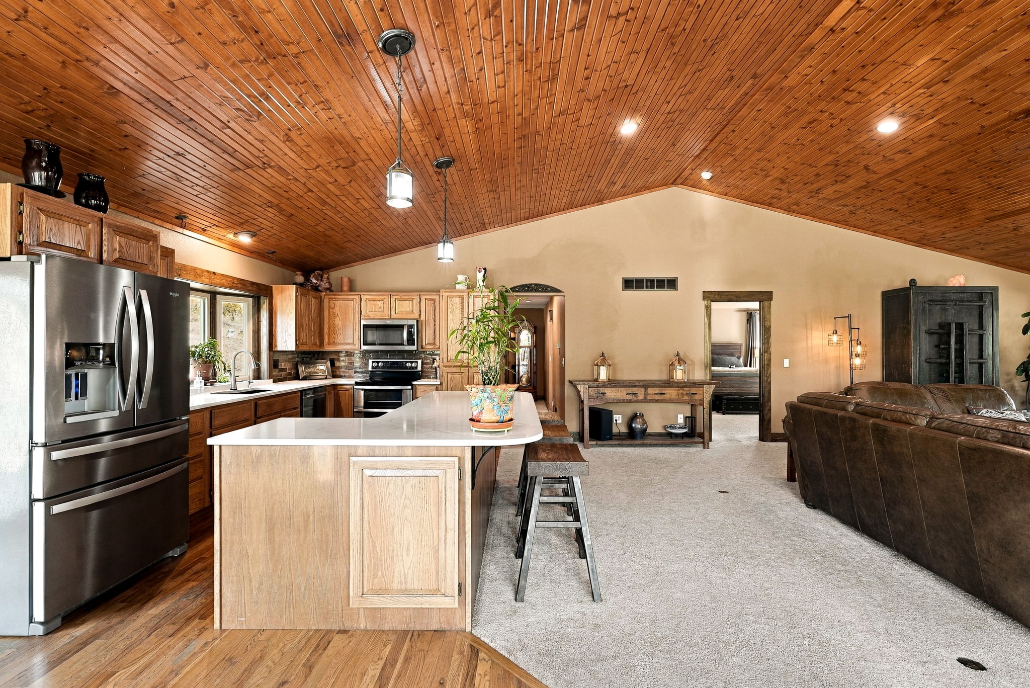 This open-concept kitchen features a large central island with a light-colored countertop and bar seating, seamlessly flowing into a carpeted living area. The space is defined by a striking vaulted ceiling with warm-toned wood paneling and recessed lighting, complemented by light wood cabinetry and stainless steel appliances. A wide doorway leads to an adjacent bedroom, creating an airy and connected floor plan that emphasizes comfort and functionality.