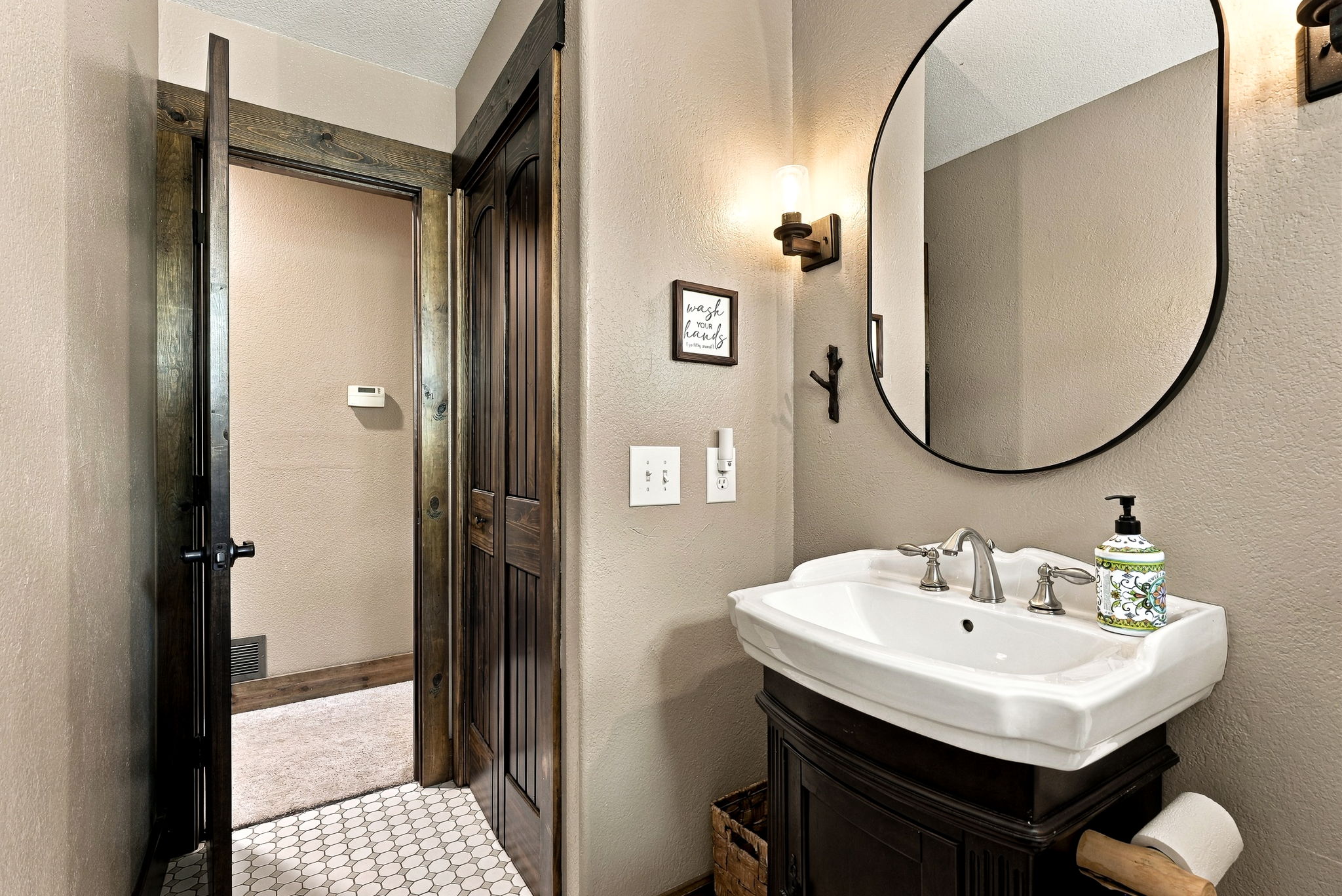 This rustic-style powder room features a dark wood vanity with a white pedestal-style sink and a decorative soap dispenser. An oval mirror hangs above the sink, complemented by a wall-mounted light fixture and a framed 'wash your hands' sign. The space includes a doorway leading to an adjacent room with carpeted flooring, while the bathroom itself is finished with a white hexagonal tile floor.