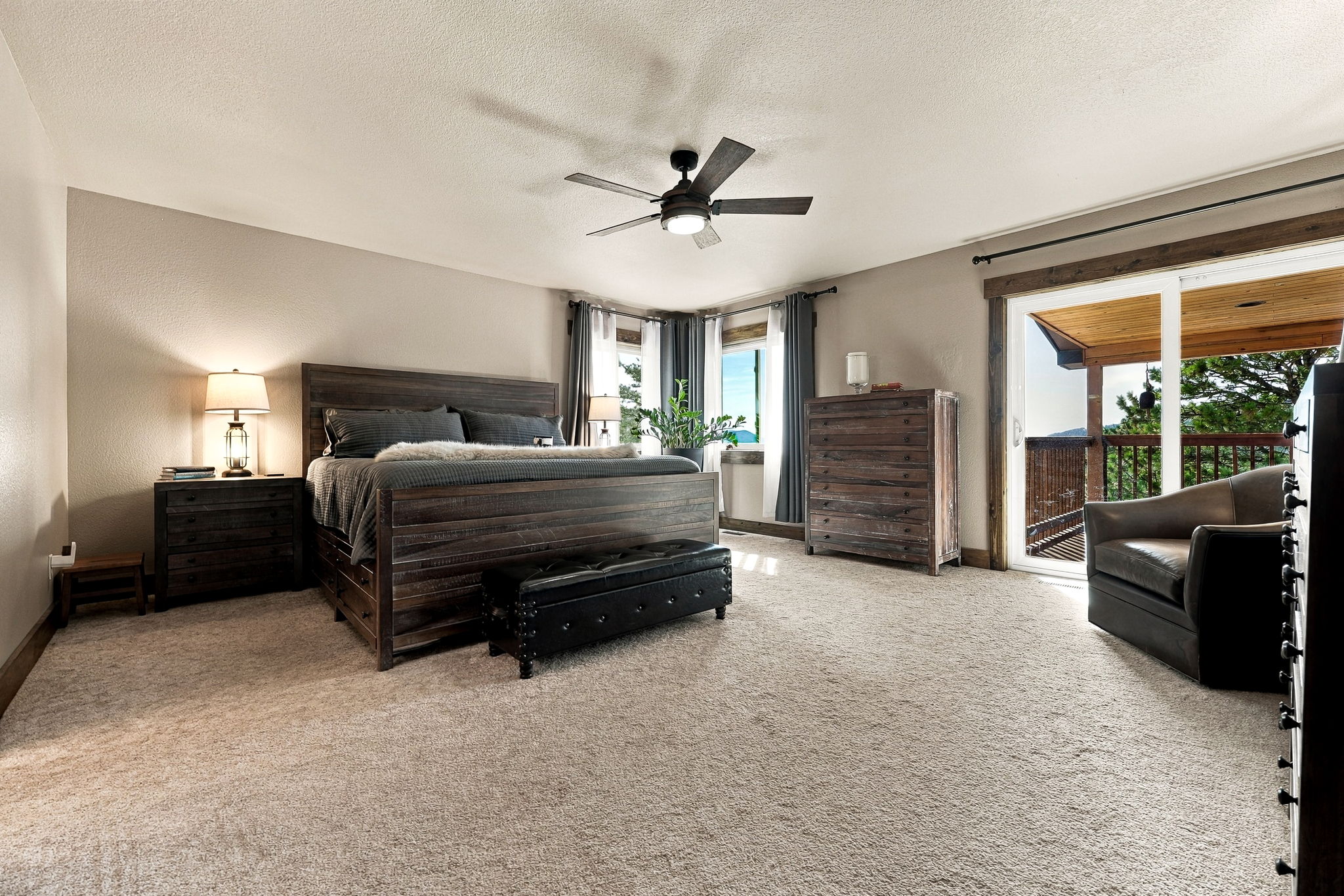 This spacious primary bedroom features a rustic aesthetic with dark wood furniture, including a substantial bed frame and matching dresser. A black leather tufted bench sits at the foot of the bed, while a comfortable leather armchair is positioned near the sliding glass doors that lead to an outdoor balcony. The room is carpeted and illuminated by a ceiling fan with integrated lighting and a bedside lamp, creating a warm and inviting atmosphere.