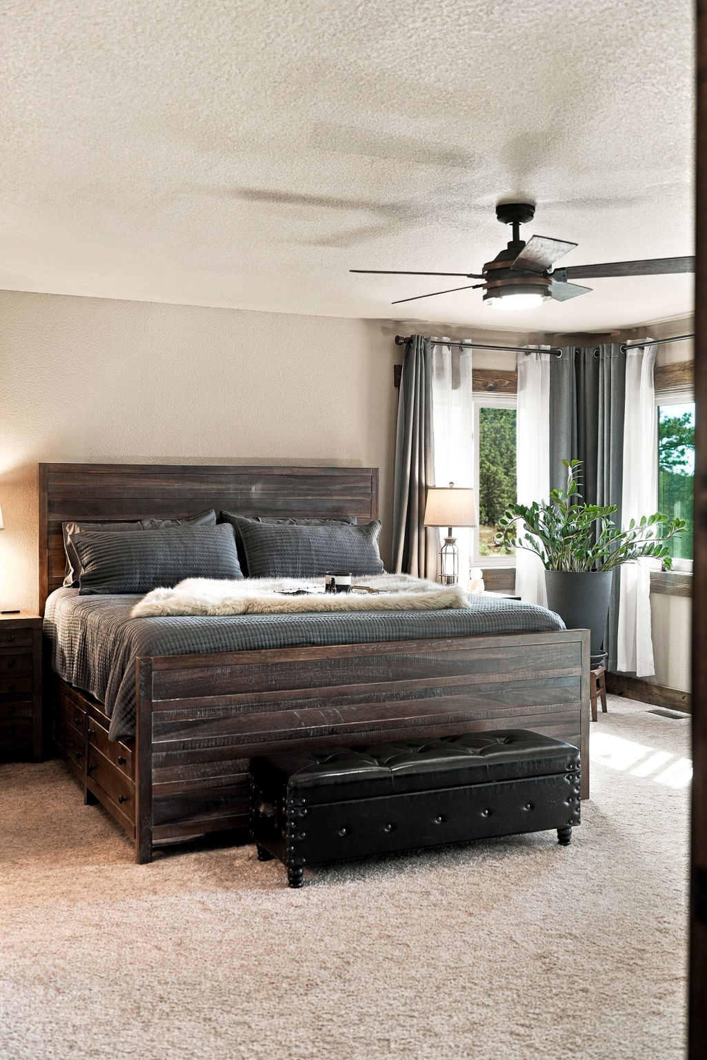This primary bedroom features a rustic, dark-stained wood bed frame with integrated storage drawers and a matching tufted leather bench at the foot. The room is accented by a ceiling fan, a large potted plant near the corner windows, and layered gray curtains that frame the outdoor view. The space is finished with neutral carpeting and warm ambient lighting, creating a cozy and inviting atmosphere.