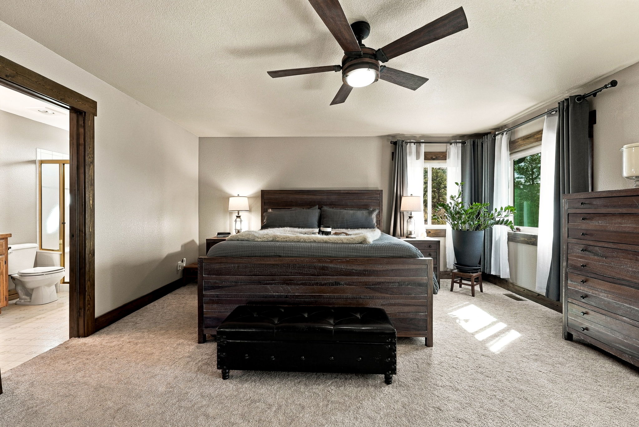 This spacious primary bedroom features a rustic aesthetic with a dark wood bed frame, matching nightstands, and a tall dresser. A black leather tufted bench sits at the foot of the bed, while a large potted plant adds a touch of greenery near the windows draped in layered curtains. The room is illuminated by a ceiling fan with integrated lighting and two bedside lamps, creating a warm and inviting atmosphere.