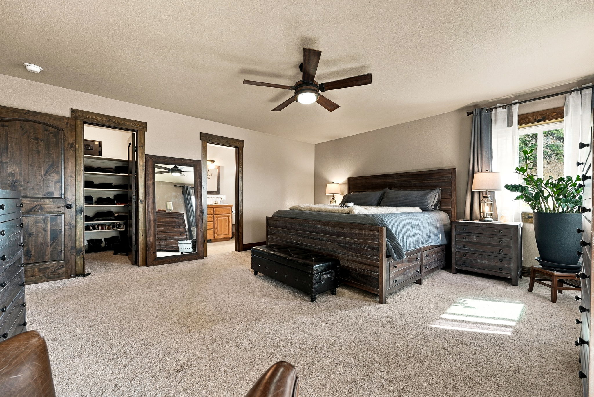 This spacious primary bedroom features a rustic aesthetic with a dark wood bed frame, matching nightstands, and a leather storage bench at the foot of the bed. The room includes a walk-in closet with built-in shelving, a full-length floor mirror, and a doorway leading to an adjoining bathroom. Large windows with layered curtains allow natural light to brighten the neutral-toned walls and carpeted floor.