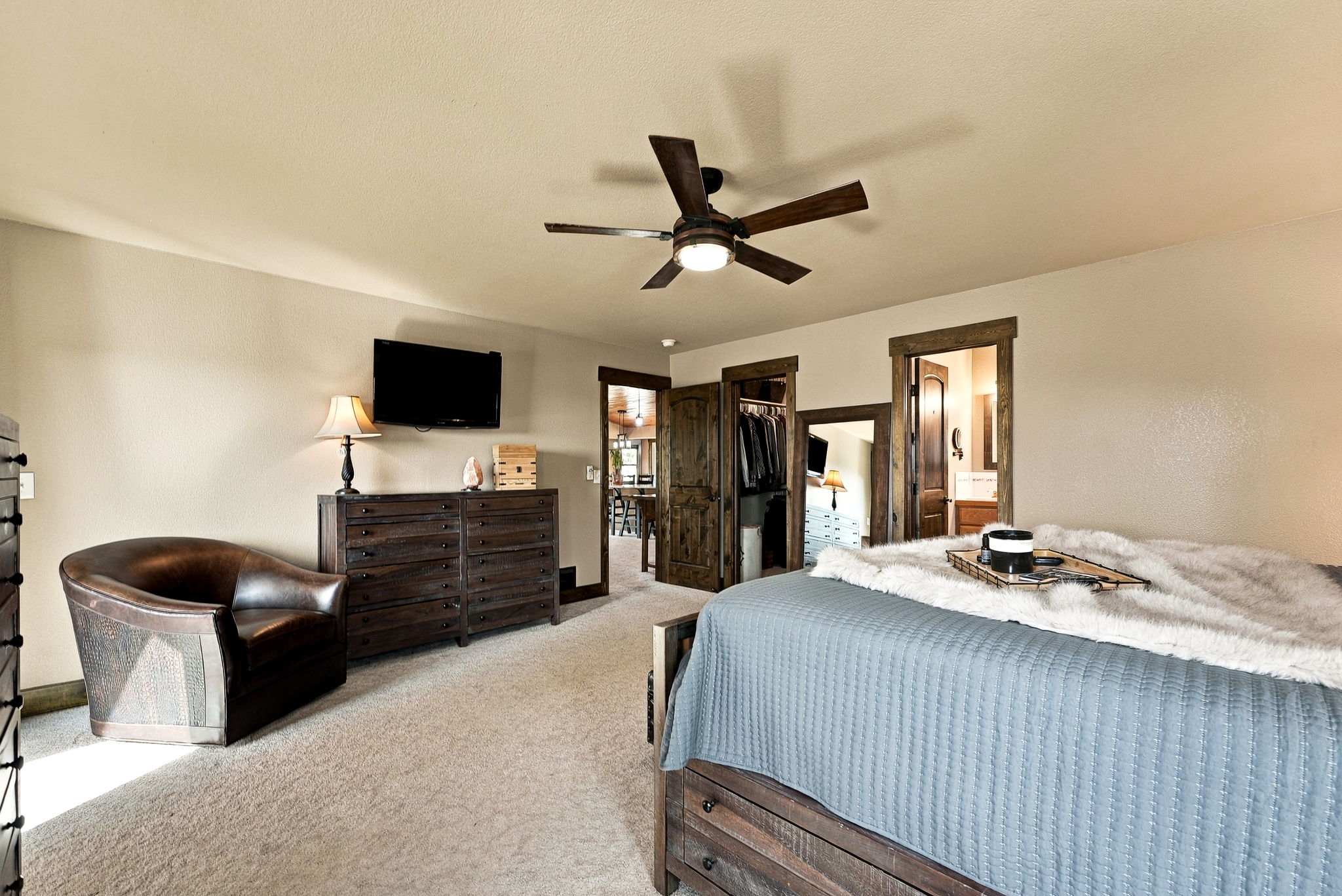 This primary bedroom features a rustic aesthetic with dark wood furniture, a plush bed topped with a textured throw, and a leather armchair. The room includes a ceiling fan, neutral carpeting, and multiple doorways leading to a walk-in closet and an adjoining bathroom. The perspective captures the cozy, well-appointed layout of the space, emphasizing its comfortable and inviting atmosphere.
