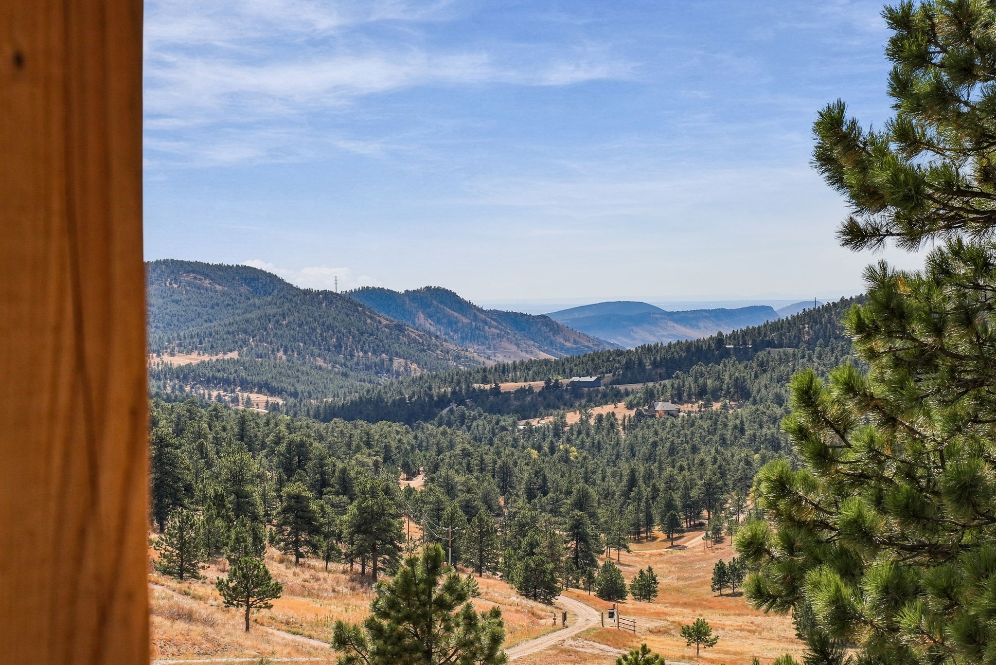 This image captures a scenic, elevated view of a mountainous landscape, framed by a wooden post on the left and pine branches on the right. The perspective suggests the view is from a balcony or deck, looking out over a valley filled with dense evergreen forests, rolling hills, and a winding dirt road. The scene conveys a sense of tranquility and seclusion, highlighting the natural beauty and expansive vistas of the property's surroundings.