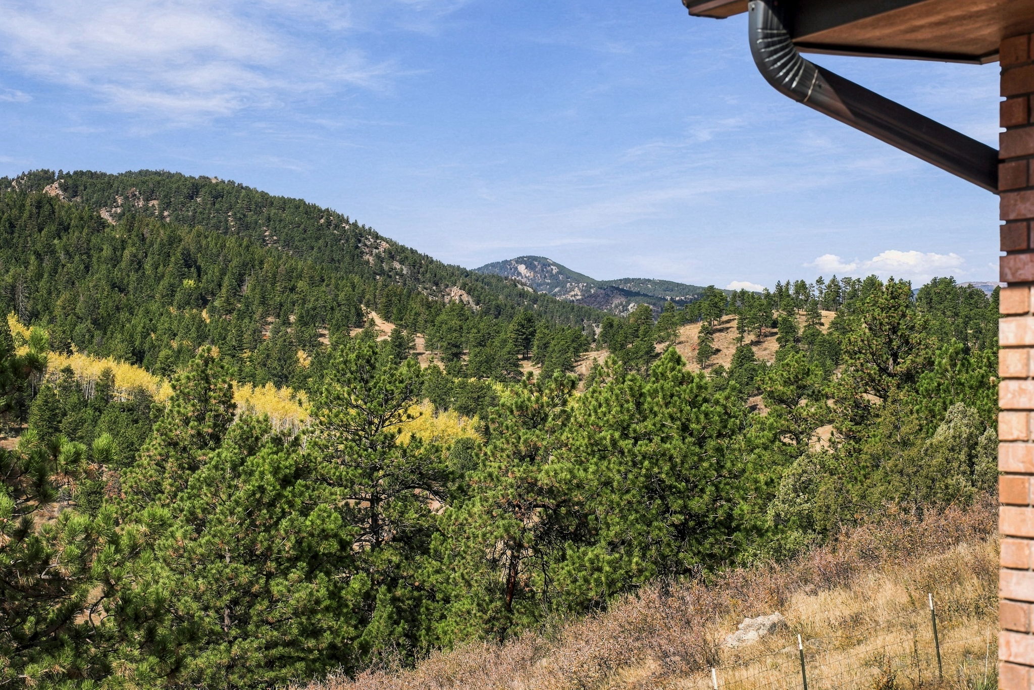 This view from an elevated deck or balcony captures a sweeping, panoramic vista of a dense, mountainous forest landscape. The foreground features a brick corner column and a metal gutter system, framing the natural scenery of evergreen trees and patches of golden-hued deciduous trees. The perspective offers a serene, high-altitude vantage point that emphasizes the property's connection to the surrounding wilderness.