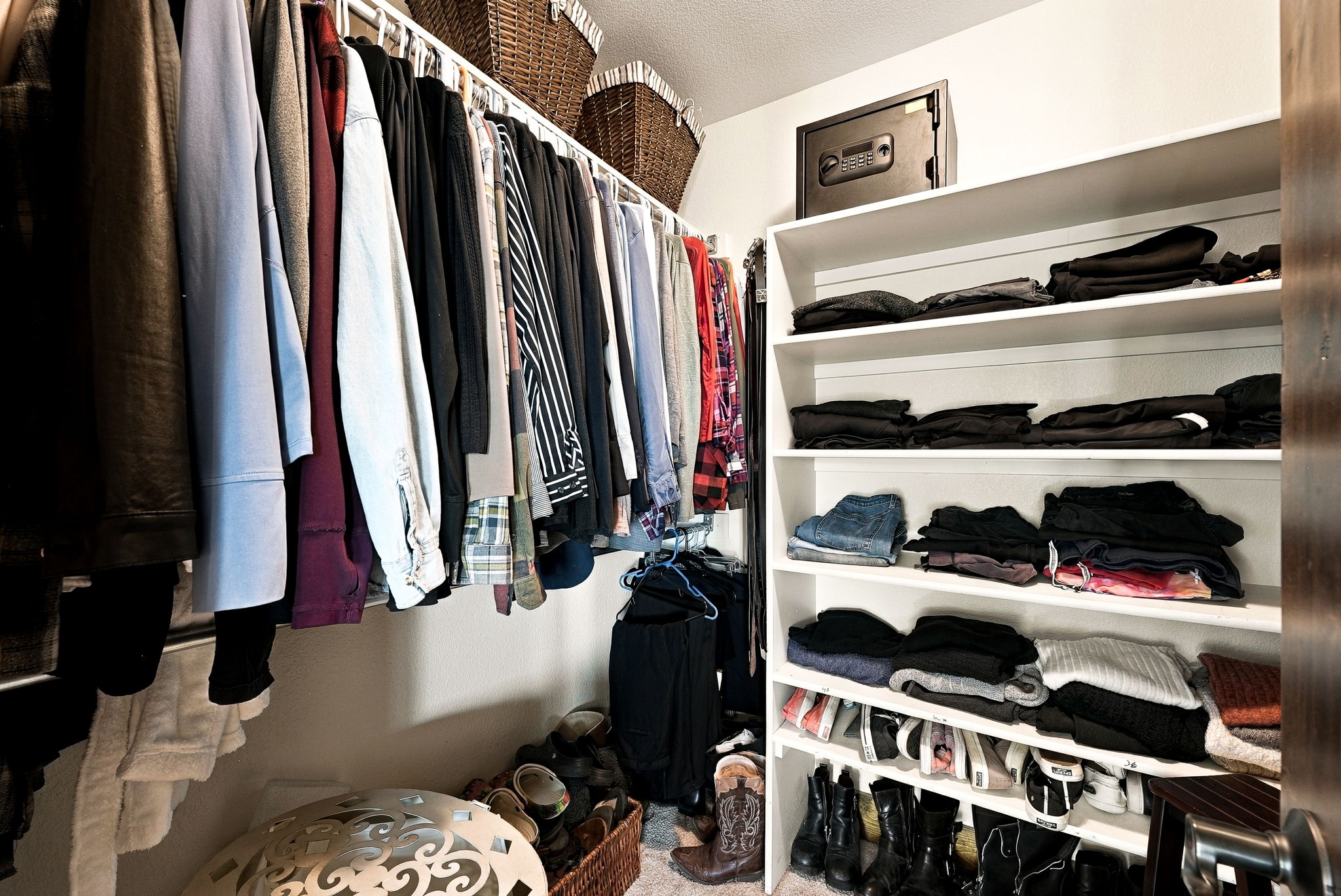 This organized walk-in closet features a combination of hanging garment rods and open shelving units. The space includes a wall-mounted safe on the top shelf, woven storage baskets, and a variety of folded clothing and footwear neatly arranged throughout. The perspective is from the entrance of the closet, showcasing the efficient use of storage space.
