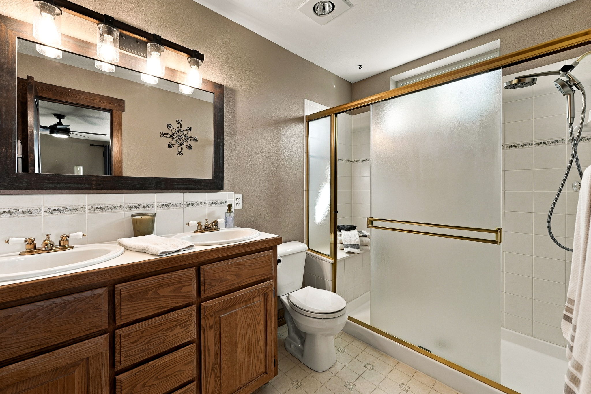 This bathroom features a dual-sink vanity with a warm-toned wood cabinet and a large mirror framed in matching wood. To the right, a walk-in shower is enclosed by sliding glass doors with a gold-toned frame and includes both a fixed showerhead and a handheld sprayer. The space is finished with neutral-toned walls, tiled flooring, and a decorative tile border above the vanity.