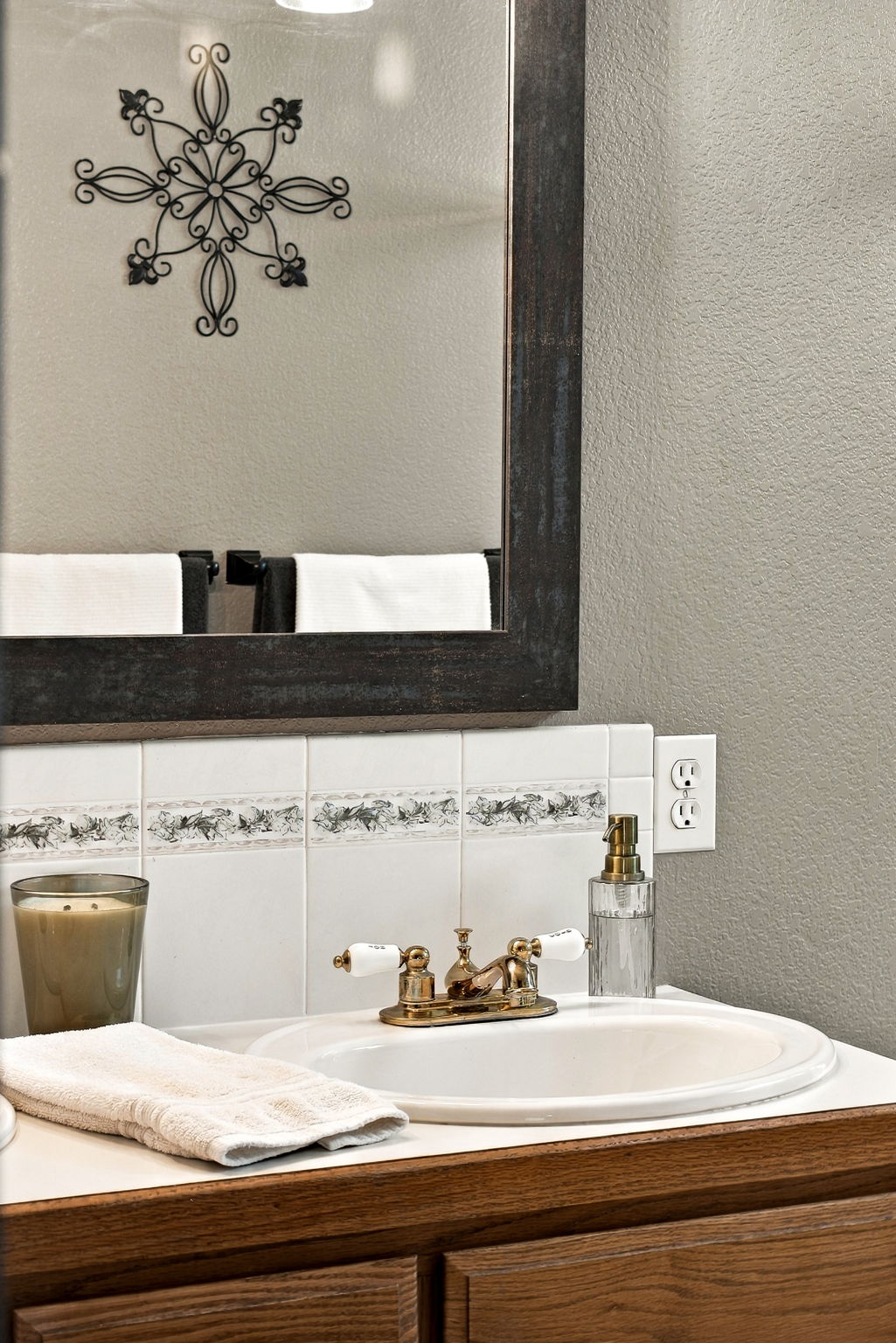 This close-up shot captures a bathroom vanity area featuring a white oval sink with a vintage-style brass faucet and white porcelain handles. The backsplash is composed of white square tiles accented with a decorative floral border, and a dark-framed mirror hangs above, reflecting a wall-mounted metal scrollwork ornament. A glass soap dispenser and a candle sit on the countertop, creating a cozy and traditional aesthetic.