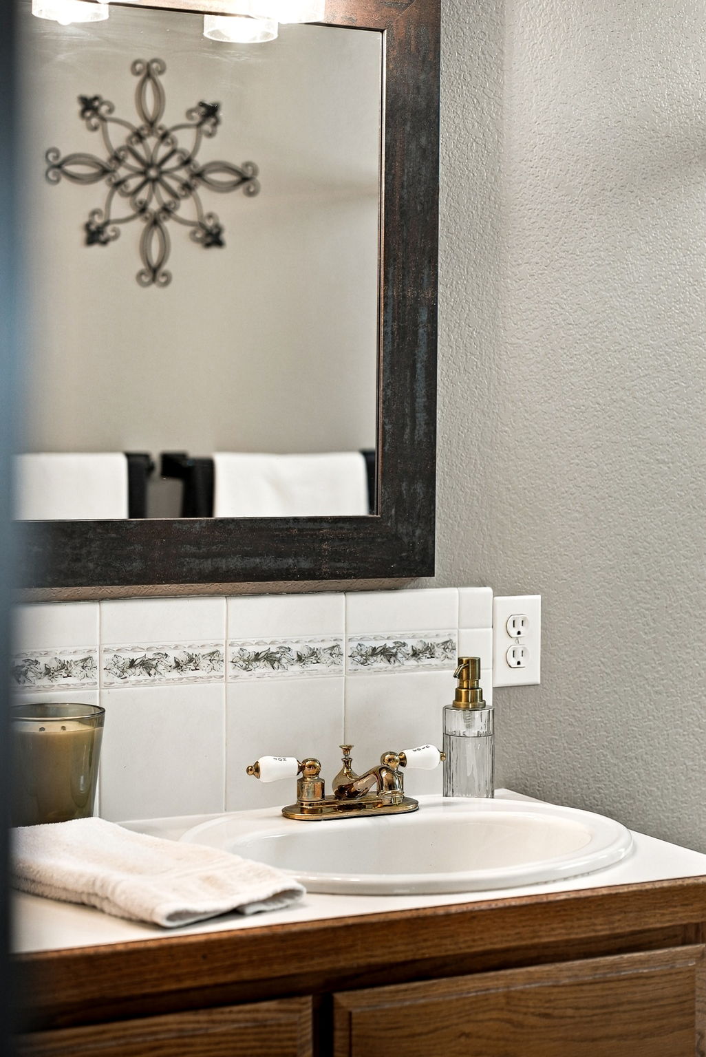This close-up shot captures a bathroom vanity featuring a white sink with a vintage-style brass faucet and white porcelain handles. The backsplash is composed of white square tiles accented with a decorative floral border, and a dark-framed mirror hangs above. A glass soap dispenser and a folded white towel add functional touches to the warm, traditional aesthetic.