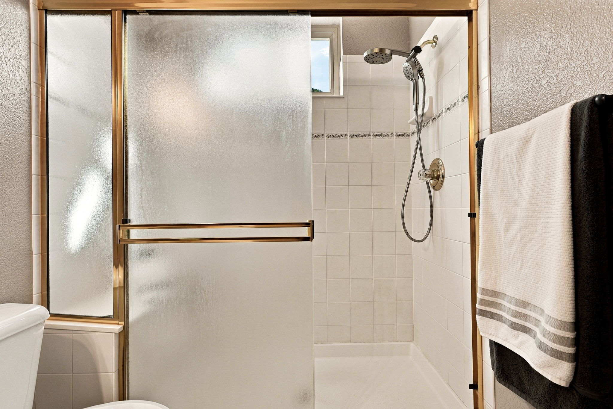 This bathroom features a walk-in shower enclosed by frosted glass sliding doors with a gold-toned frame. The shower interior is finished with white square wall tiles, a decorative border accent, and a handheld showerhead fixture. A towel hangs on a rack to the right, and a small window provides natural light into the shower stall.