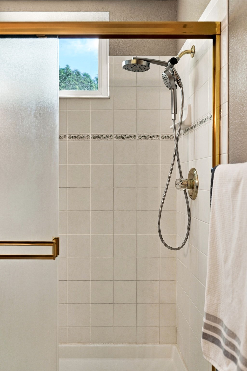 This shower stall features light-colored square wall tiles accented by a decorative floral border. It is equipped with a dual-head shower system, including a handheld sprayer, and is enclosed by a sliding glass door with a gold-toned frame. A window above the shower allows natural light to enter the space, while a towel hangs on the right side.