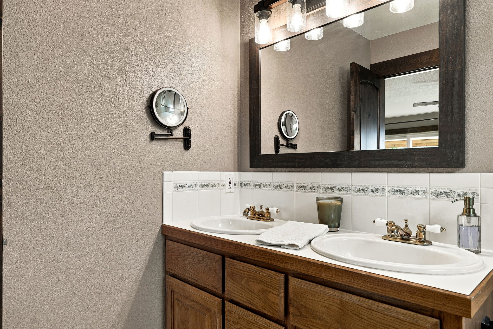 This double-vanity bathroom features a warm wood cabinet base topped with a white counter and two inset sinks. A large, dark-framed mirror hangs above the vanity, complemented by a rustic-style light fixture with multiple glass shades. The wall is finished with a neutral textured paint and a decorative tile backsplash, creating a cozy and functional space.