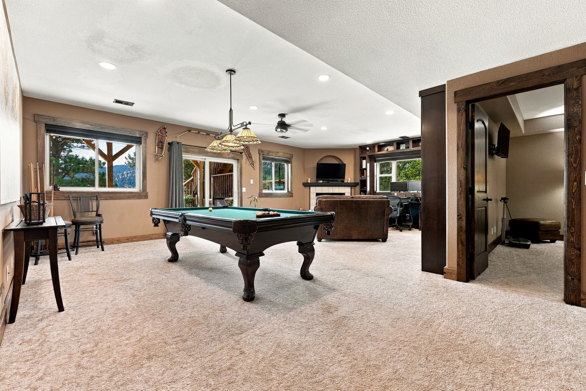 This spacious basement recreation room features a central pool table illuminated by a multi-light pendant fixture. The room includes a cozy seating area with a leather sofa facing a television mounted above a fireplace, alongside a dedicated office nook. The space is finished with neutral carpeting, warm-toned walls, and dark wood trim, creating a comfortable and inviting atmosphere for entertainment.