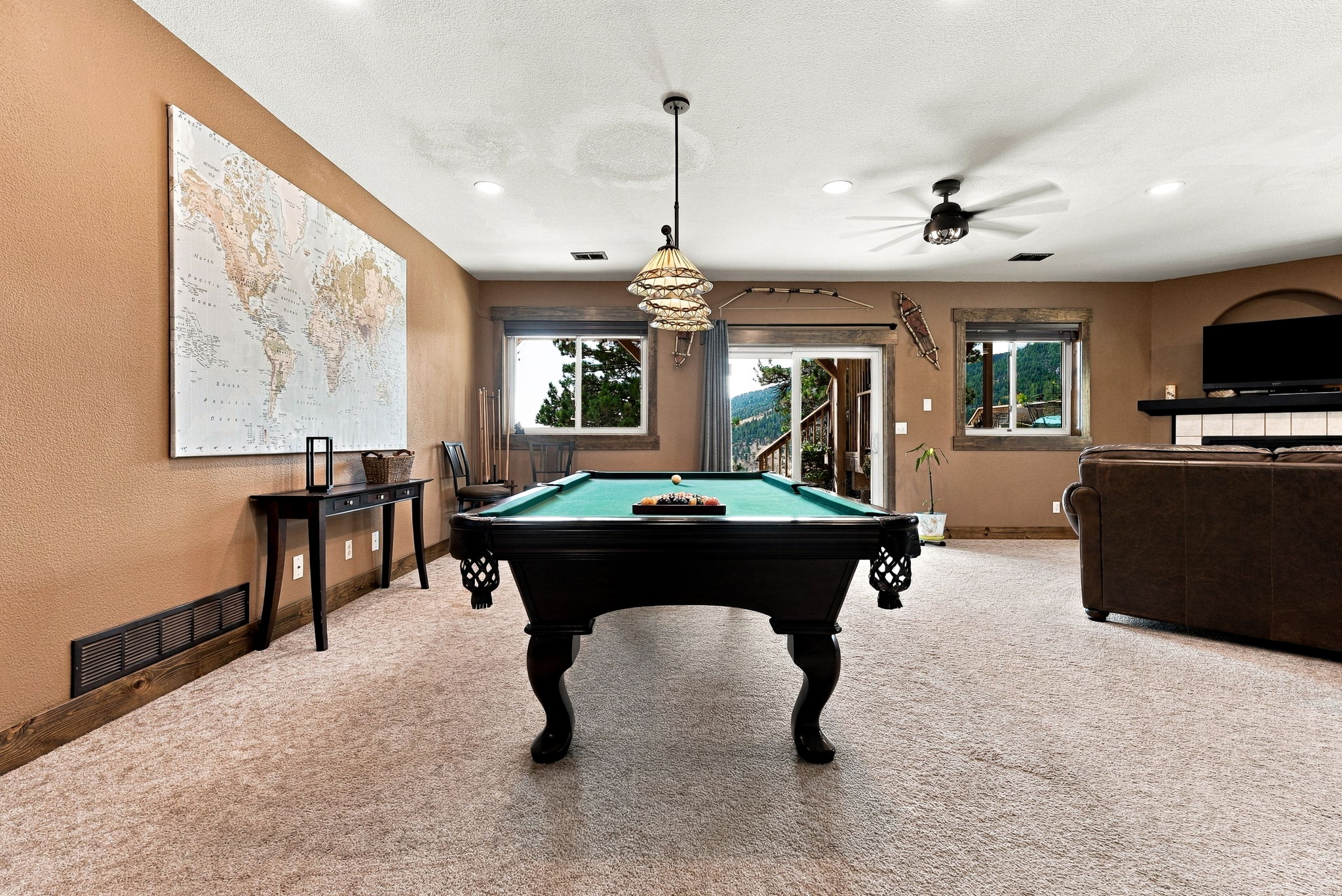 This spacious basement recreation room features a central pool table as the primary focal point, set against a backdrop of warm-toned walls and neutral carpeting. The room includes a large world map wall art, a console table, and a comfortable seating area with a television mounted above a fireplace. Large windows and a sliding glass door provide natural light and views of the surrounding landscape, creating an inviting space for entertainment.