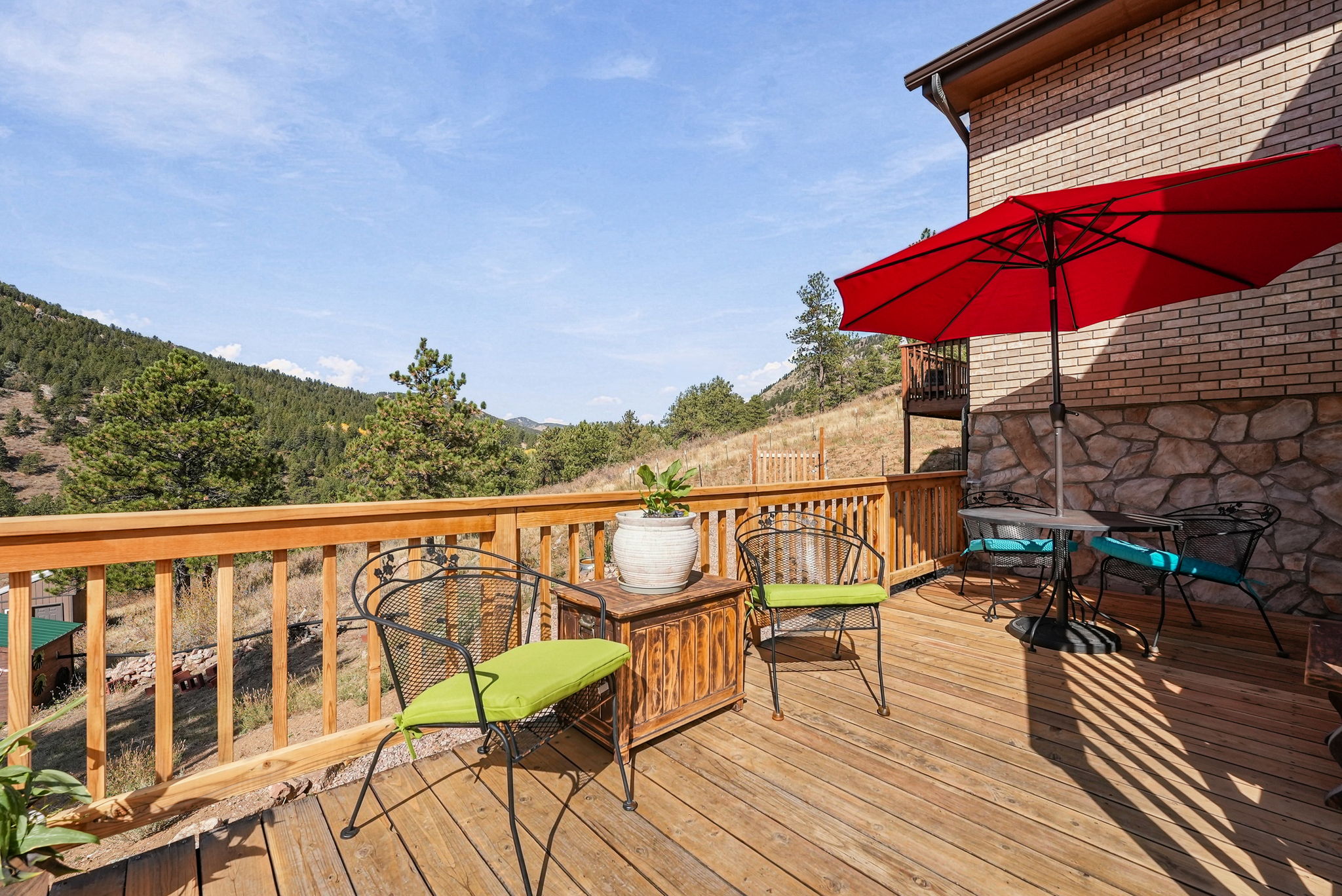 This elevated wooden deck offers a scenic outdoor living space with a vibrant red umbrella providing shade over a small bistro set. The deck features a sturdy wooden railing and is furnished with additional chairs and a small side table topped with a potted plant, all set against a backdrop of rolling hills and forest. The perspective captures the inviting atmosphere of the space, highlighting the expansive mountain views and the warm, natural wood tones of the deck.