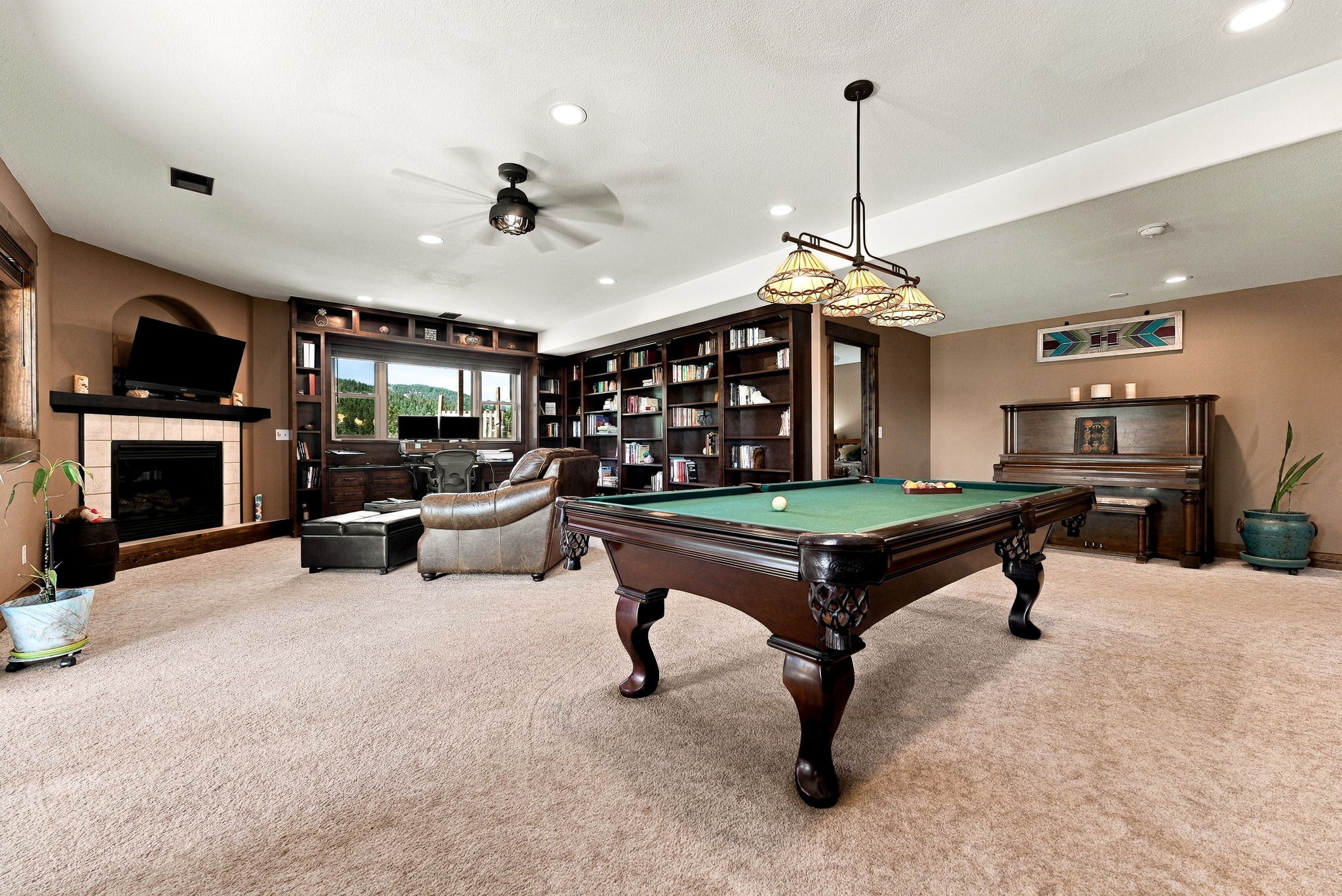 This spacious basement recreation room features a large pool table as the central focal point, illuminated by a multi-light pendant fixture. The room includes a dedicated office area with built-in dark wood shelving, a cozy fireplace with a television mounted above, and an upright piano against the far wall. The space is finished with neutral carpeting and warm-toned walls, creating a comfortable and versatile living area.