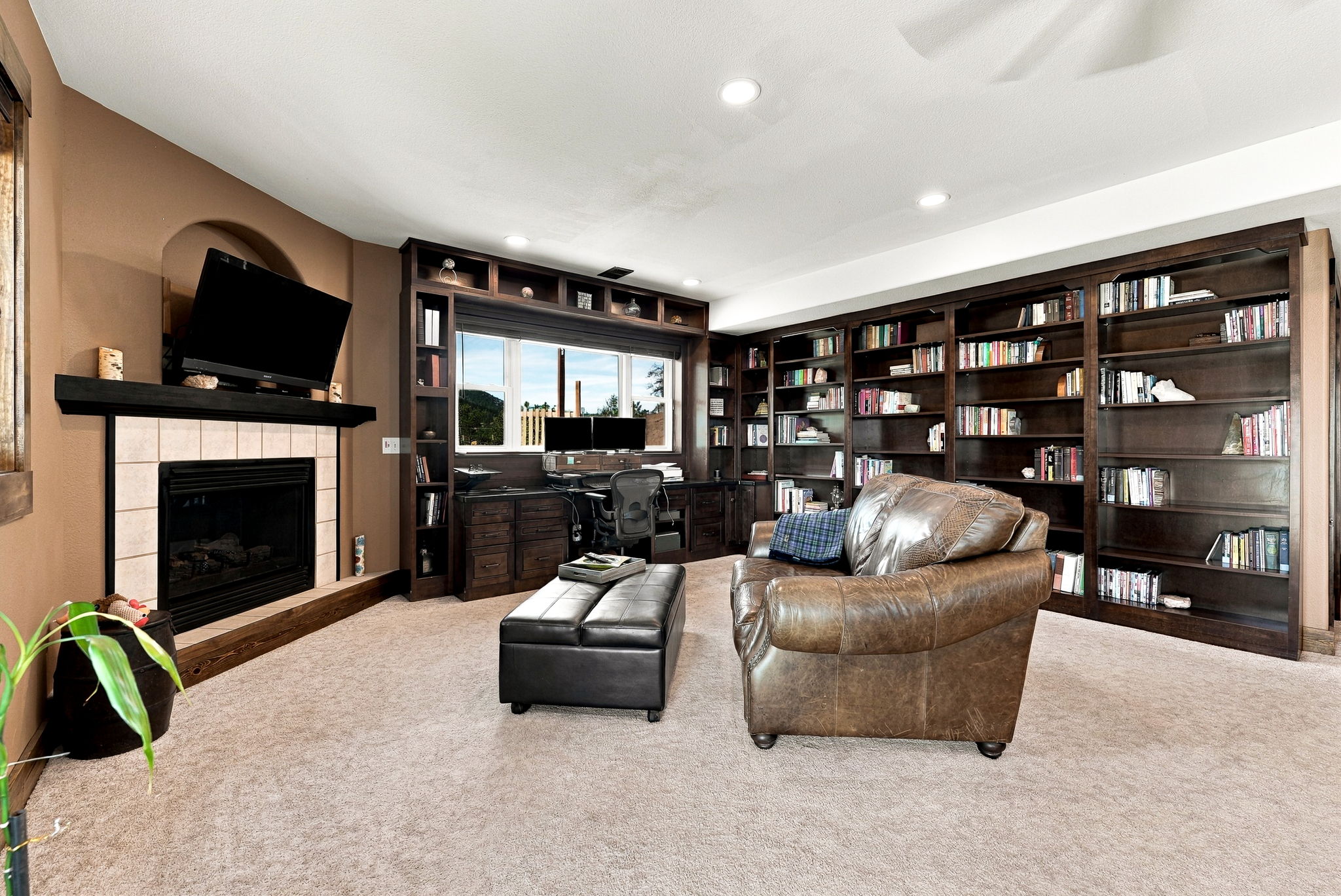 This spacious home office and library features extensive dark wood built-in shelving that spans the wall, providing ample storage and display space. A central desk area is positioned beneath a window, while a comfortable leather armchair and a matching ottoman create a cozy reading nook. To the left, a fireplace with a dark mantel and a television mounted above serves as a focal point, completing this sophisticated and functional multi-purpose room.