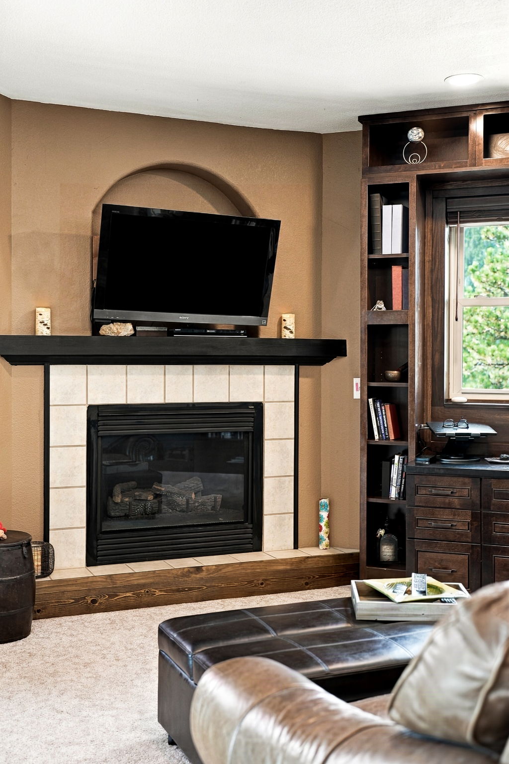 This living room features a cozy fireplace with a light-colored tile surround and a dark wood mantel, topped by a wall-mounted television set within a recessed arched niche. To the right, a built-in dark wood shelving unit provides ample storage and display space, integrated with a desk area near a window. The room is anchored by a dark leather ottoman in the foreground, creating a warm and inviting atmosphere.