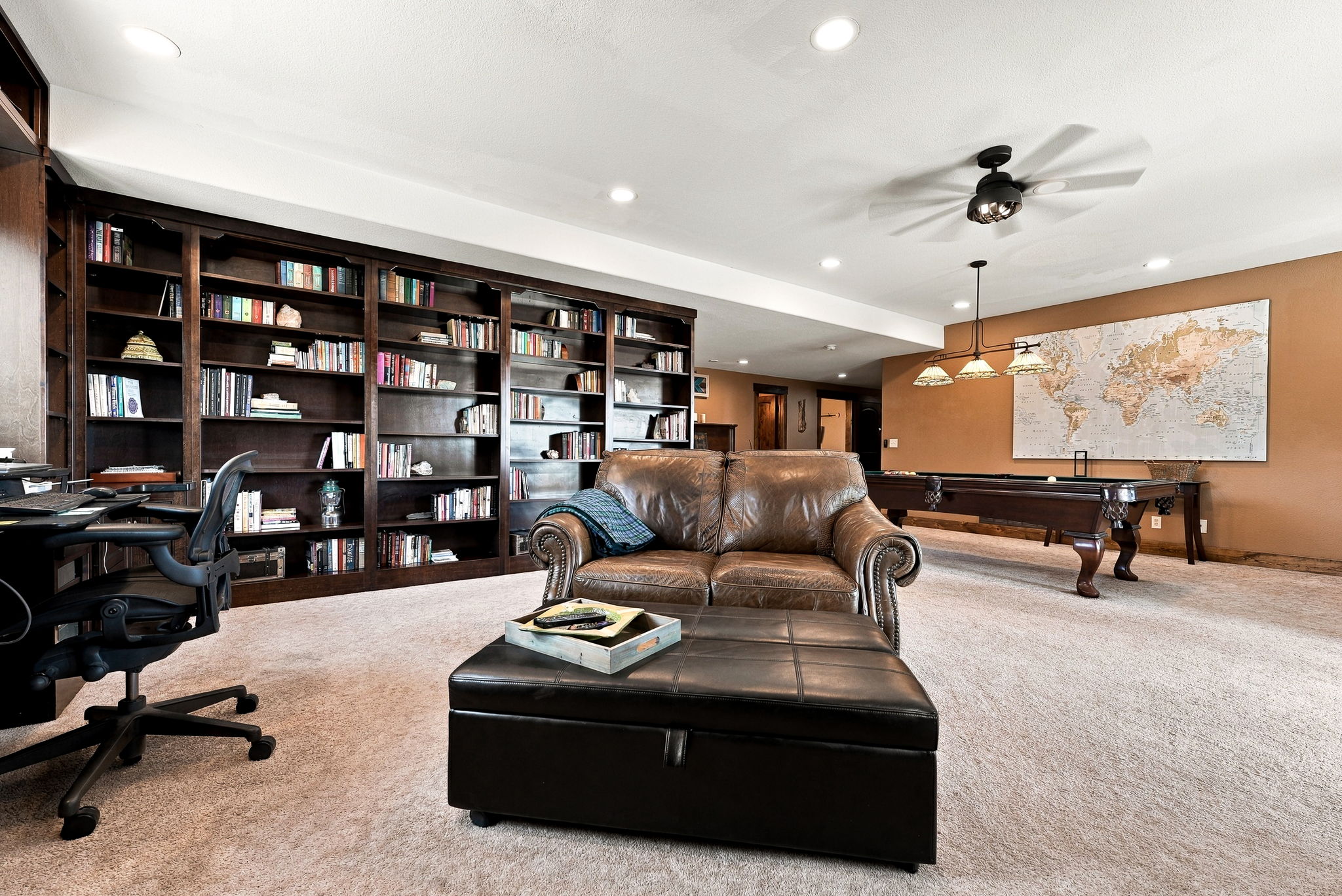 This spacious finished basement features a versatile layout that combines a cozy seating area with a dedicated home office and a game room. A large, dark wood built-in bookshelf spans the wall, providing ample storage, while a leather loveseat and matching ottoman create a comfortable lounge space. In the background, a pool table sits beneath a classic billiard light, set against a warm-toned wall adorned with a large world map.