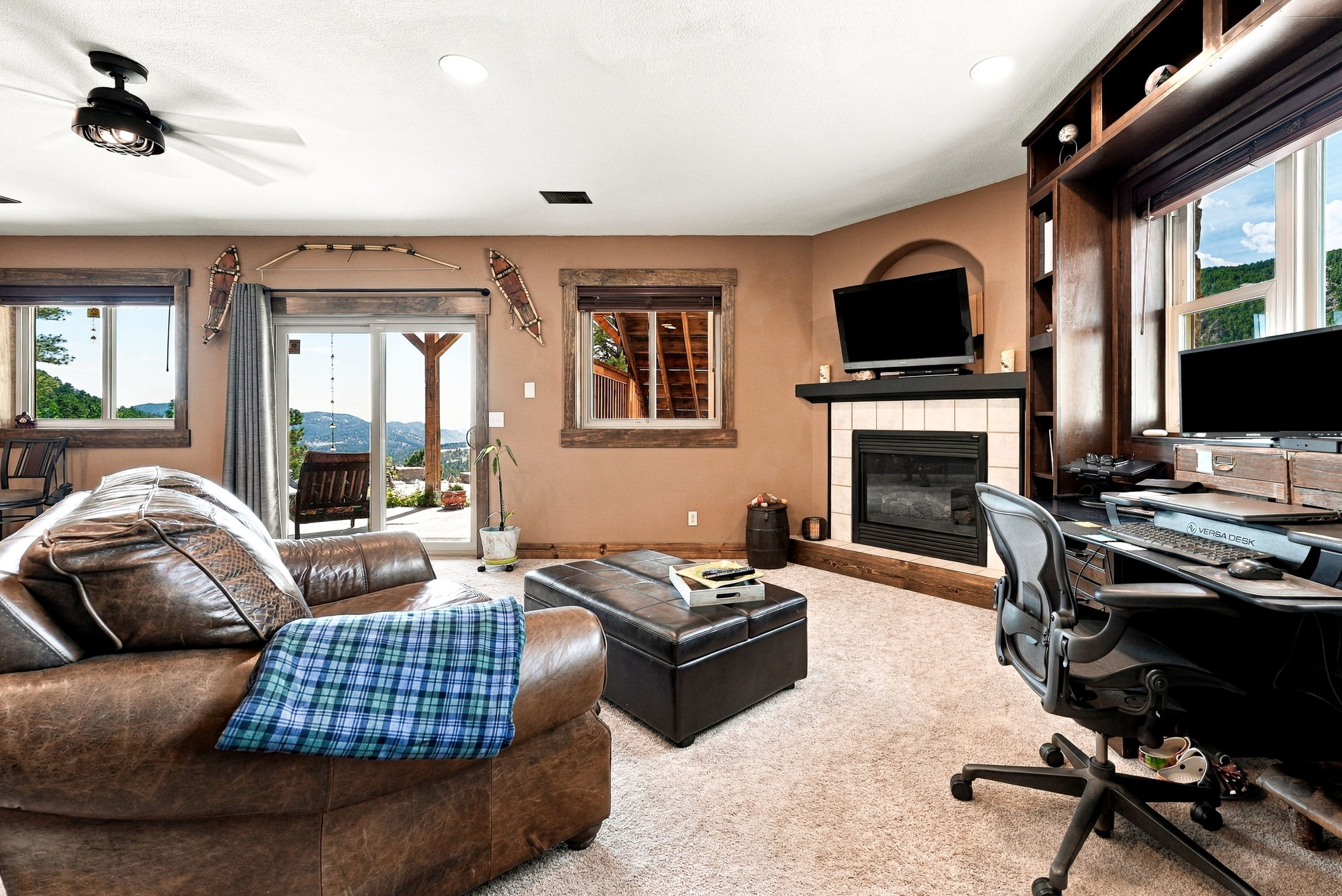 This cozy living room features a warm, rustic aesthetic with brown walls, a leather sofa, and a central ottoman. A fireplace with a tiled surround and a mounted television serves as a focal point, while a built-in wooden desk area provides a functional workspace. Large sliding glass doors open to a scenic outdoor view, filling the space with natural light and connecting the interior to the surrounding landscape.