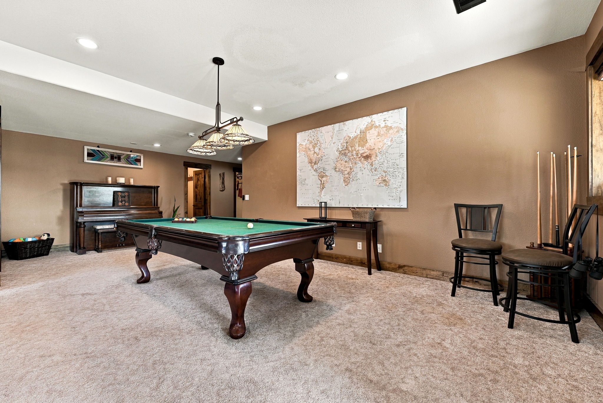 This spacious basement recreation room features a classic pool table as its centerpiece, illuminated by a multi-light pendant fixture. The room is finished with neutral carpeting and warm-toned walls, accented by a large world map wall art and a traditional upright piano. Two bar-style chairs and a cue rack are positioned to the side, creating a comfortable and inviting space for entertainment.