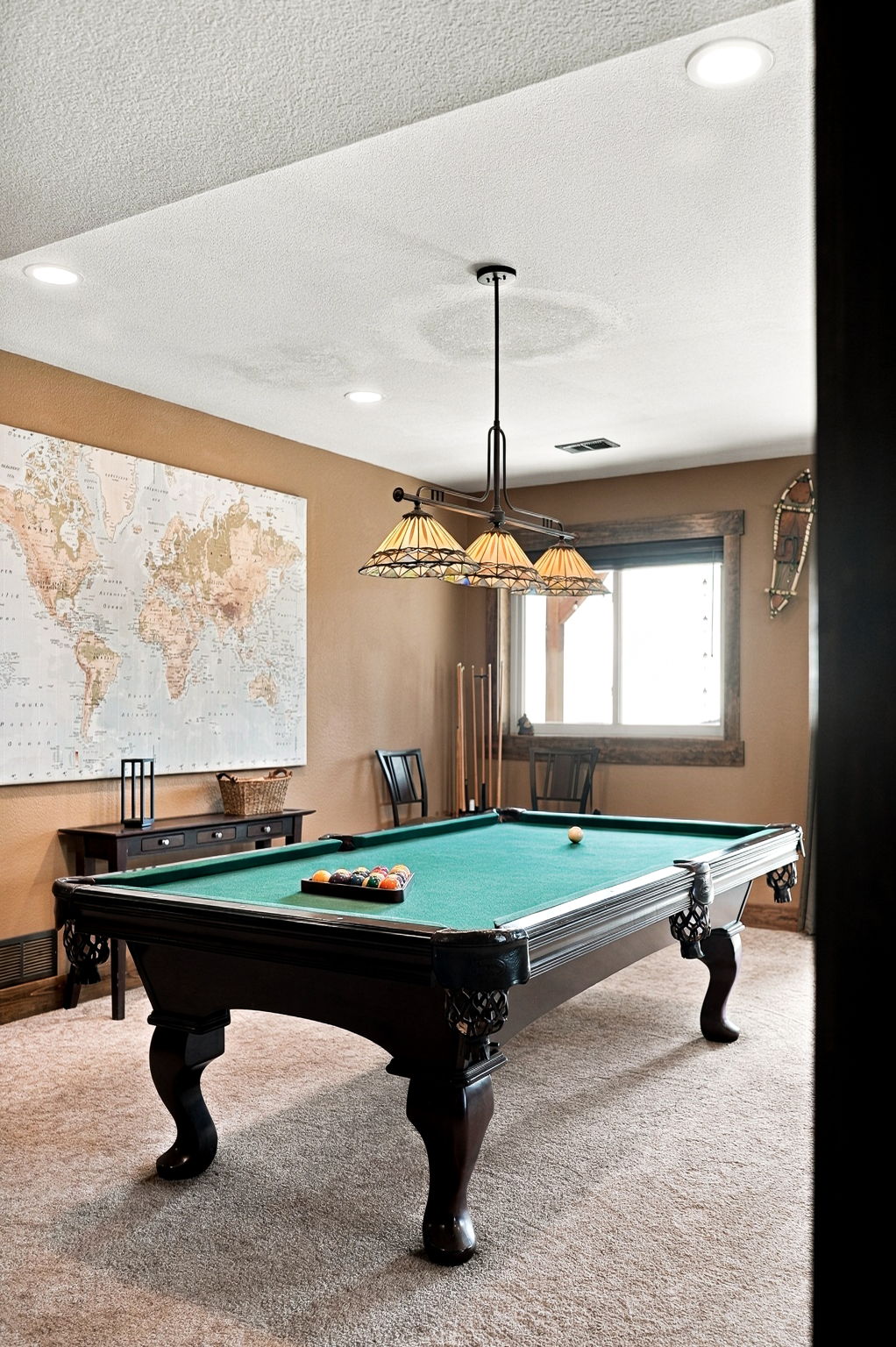 This inviting game room features a classic pool table as the central focal point, illuminated by a multi-light pendant fixture. The walls are painted in a warm, neutral tone and adorned with a large world map, while a window with rustic wood trim allows natural light to enter. The space is carpeted and includes a console table and chairs, creating a comfortable area for recreation.