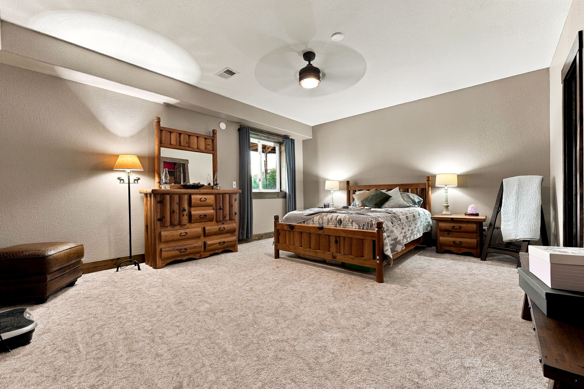 This spacious primary bedroom features a rustic aesthetic with matching light wood furniture, including a bed frame, dresser with mirror, and nightstand. The room is carpeted in a neutral tone and includes a leather ottoman, a blanket ladder, and a ceiling fan for climate control. A window with dark curtains allows natural light to enter, creating a warm and inviting atmosphere.