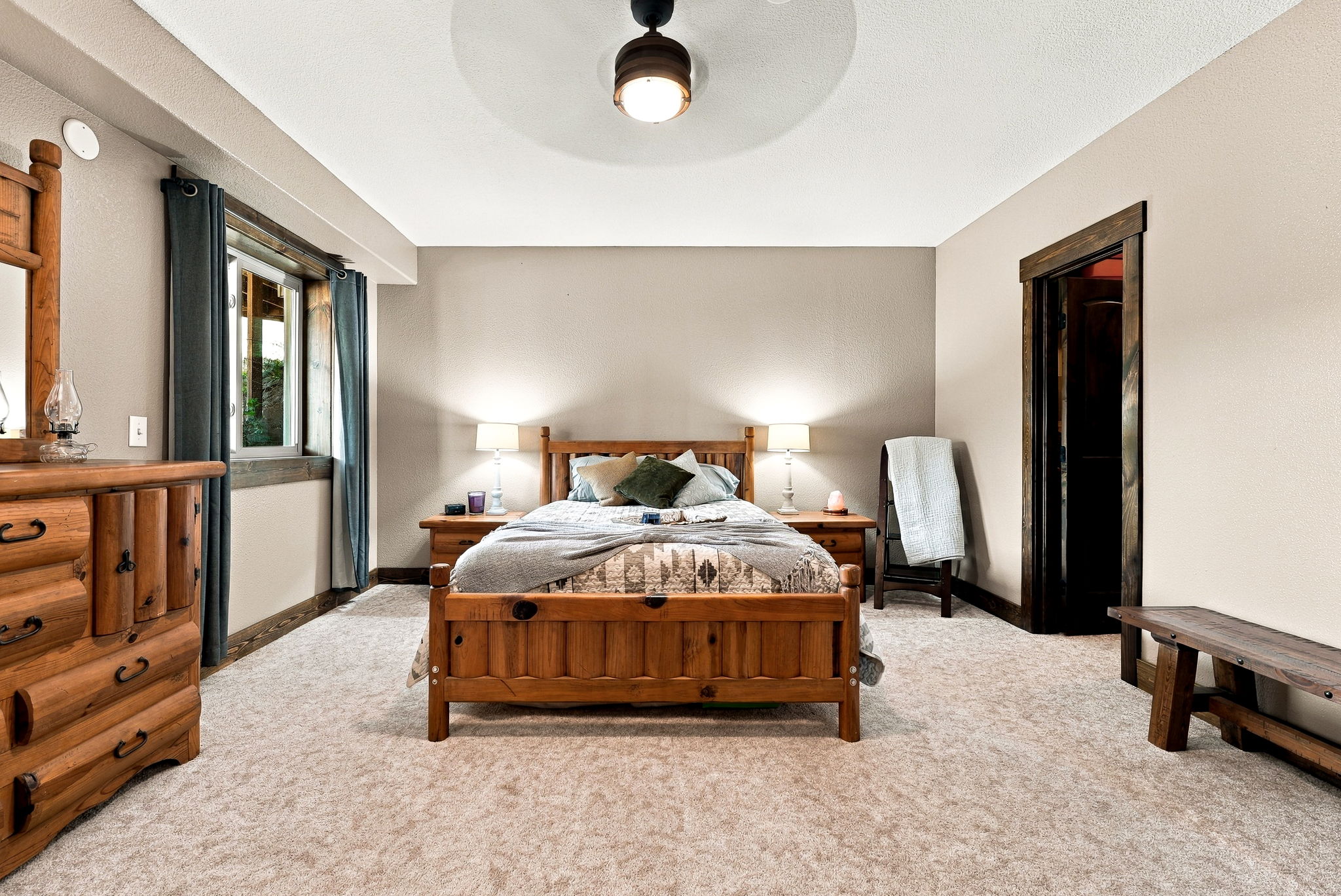 This cozy bedroom features a rustic wooden bed frame as the central focal point, flanked by matching nightstands and lamps. The room is carpeted in a neutral tone and includes a large wooden dresser on the left, a wooden bench on the right, and a window dressed with dark curtains. The space is illuminated by a ceiling fan with an integrated light fixture, creating a warm and inviting atmosphere.
