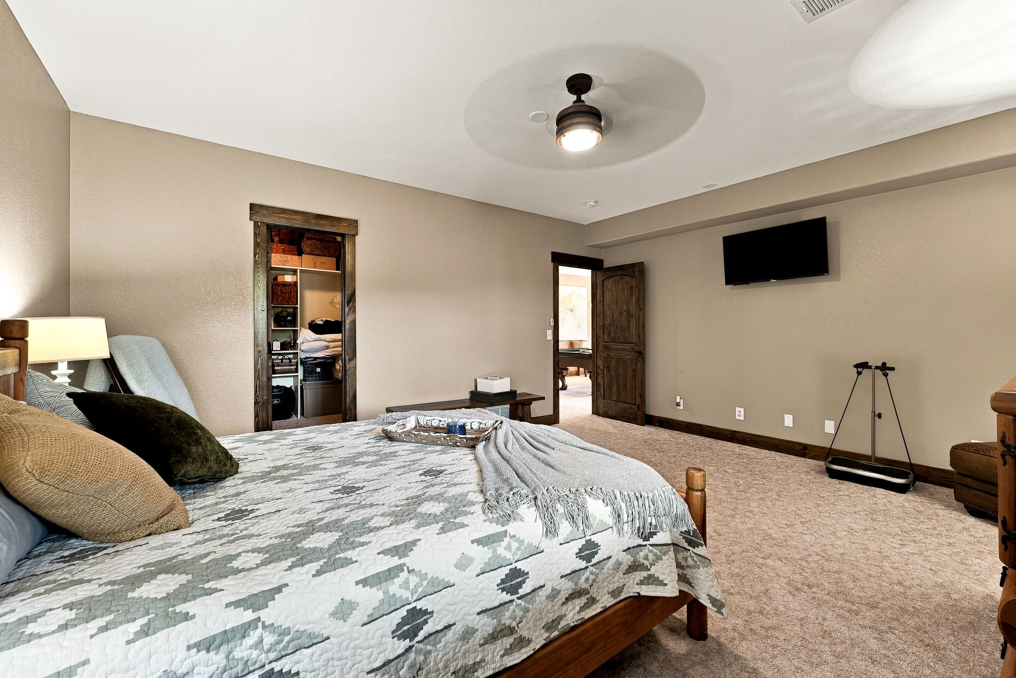 This spacious bedroom features neutral-toned walls, plush carpeting, and a wooden bed frame dressed in a patterned quilt. A ceiling fan provides overhead lighting, while a wall-mounted television and a doorway leading to an adjacent room create a functional layout. The room includes a walk-in closet with open shelving and a bench, offering a comfortable and well-organized living space.