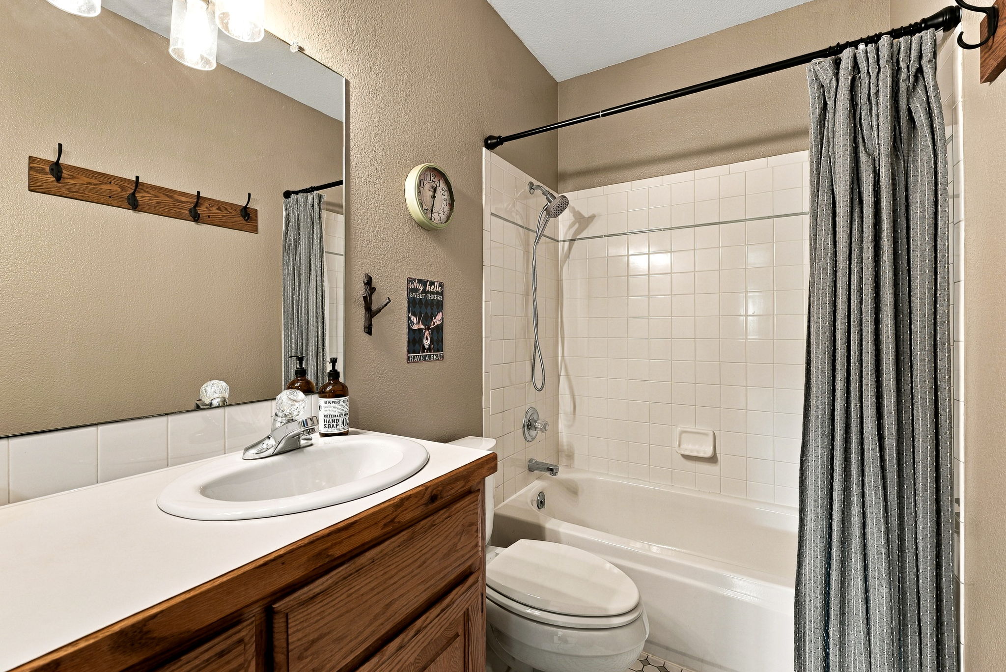 This compact bathroom features a wood vanity with a white countertop and an integrated sink, paired with a large wall-mounted mirror. A shower-tub combination is enclosed by a patterned curtain and features white wall tiles. The space is decorated with a wall clock and whimsical wall art, creating a functional and cozy atmosphere.