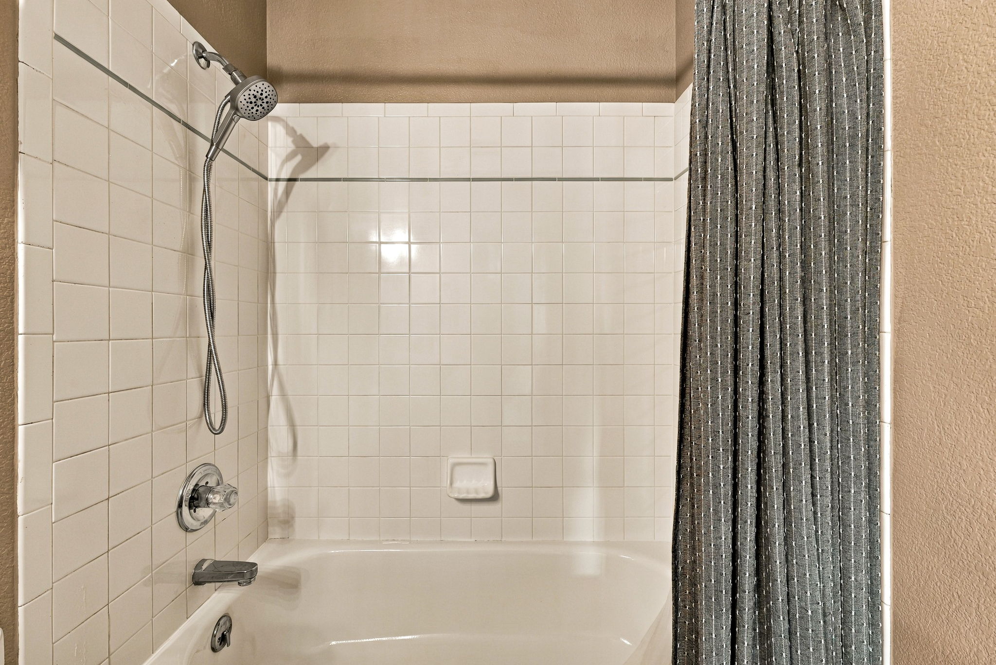 This bathroom features a standard tub-shower combination with white square wall tiles and a contrasting dark gray shower curtain. The setup includes a chrome handheld showerhead, a matching faucet, and a built-in soap dish integrated into the tiled wall. The neutral beige wall paint above the tile adds a warm tone to the functional space.