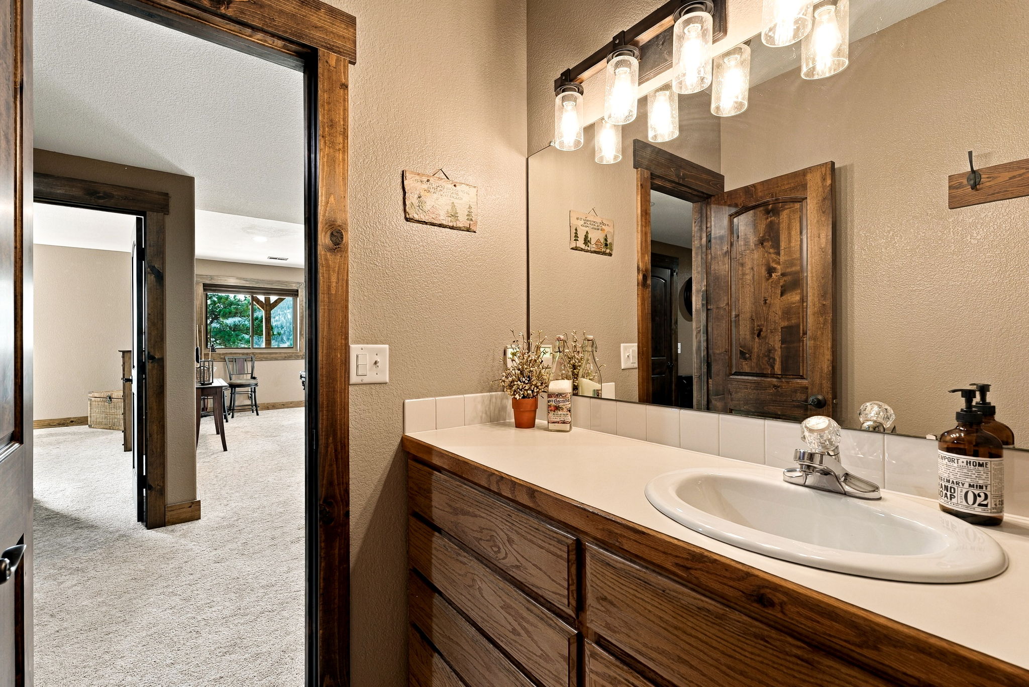This bathroom features a rustic aesthetic with a wood-framed vanity and a matching mirror frame. The space includes a white countertop with an integrated sink, a tiled backsplash, and a multi-light fixture with glass shades. A doorway provides a view into an adjacent carpeted room, creating a sense of flow between the bathroom and the rest of the home.