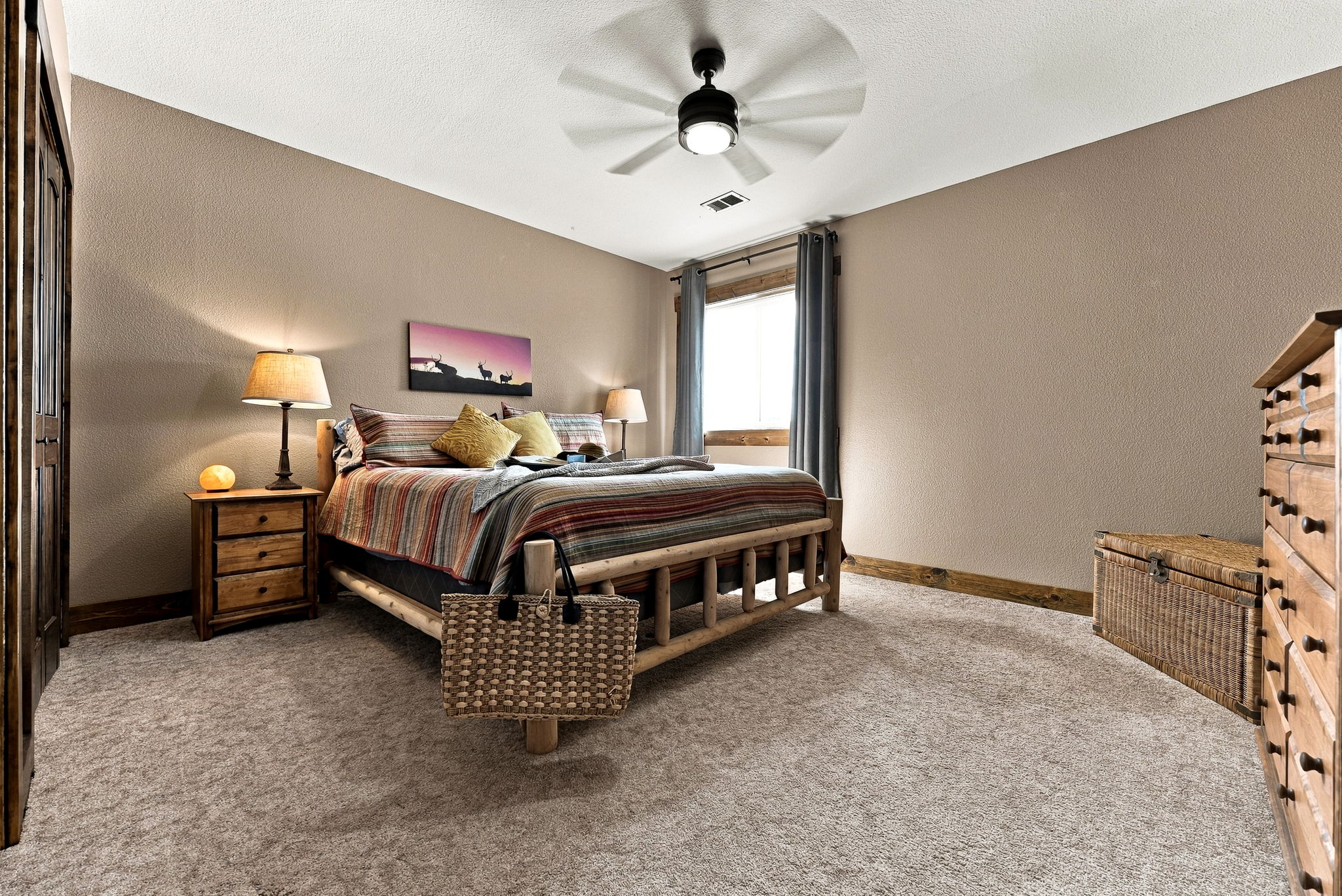 This cozy bedroom features a rustic log-style bed frame dressed in warm, striped bedding and accented with yellow throw pillows. The room is furnished with a matching wooden nightstand, a wicker storage chest, and a tall dresser, all set against neutral-toned walls and carpeted flooring. A ceiling fan with integrated lighting provides overhead illumination, while a window framed by dark curtains allows natural light to enter the space.