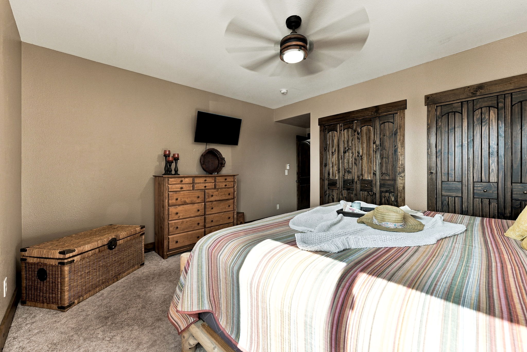This cozy bedroom features a rustic aesthetic with dark wood-paneled closet doors and a matching wooden dresser. A large bed with a striped quilt is the focal point, accented by a straw hat and a light-colored throw blanket. The room is completed by a woven storage chest at the foot of the bed and a ceiling fan, creating a warm and inviting atmosphere.