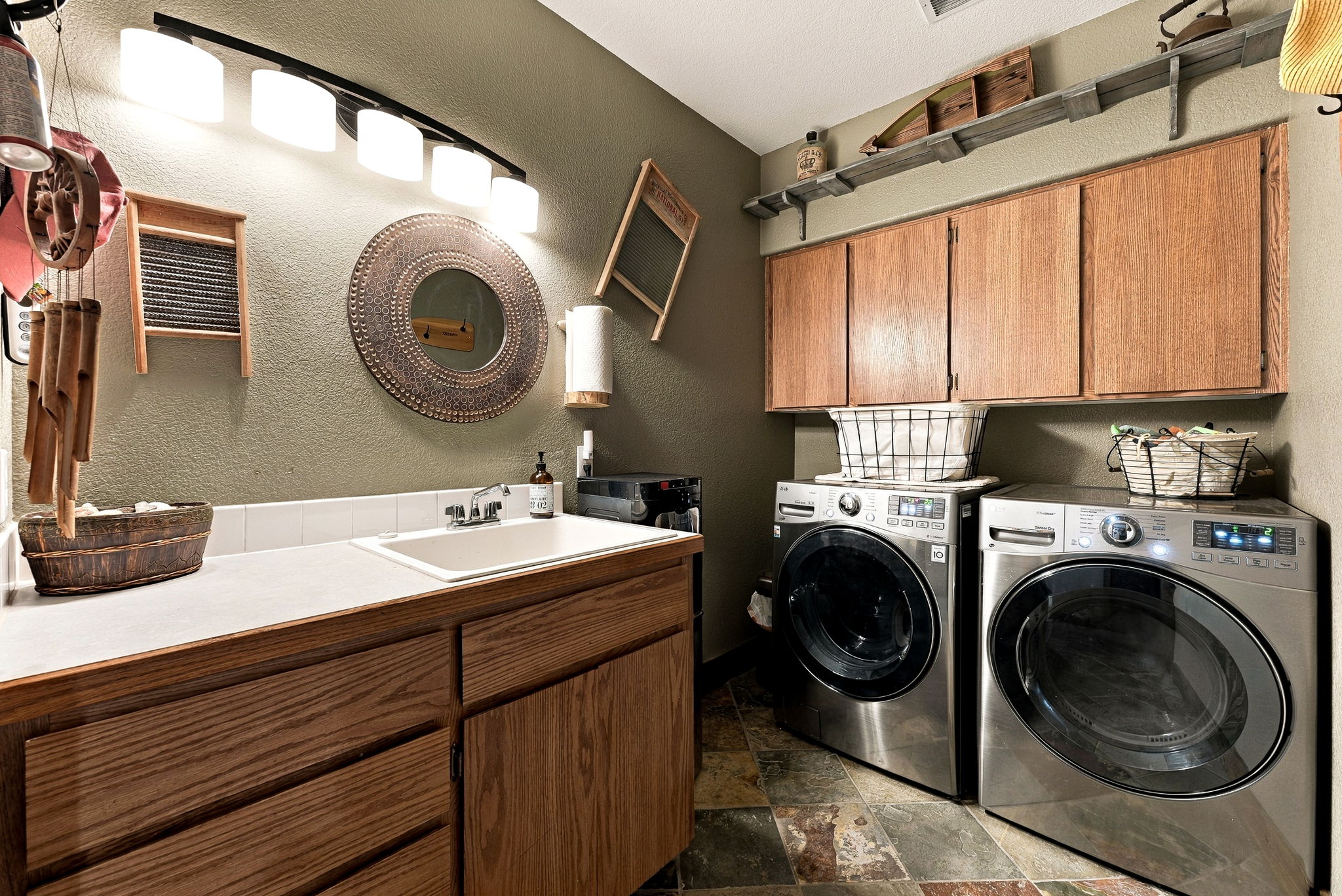 This functional laundry room features a utility sink set into a light wood vanity with a white countertop, complemented by matching upper cabinets for storage. The space is equipped with a modern stainless steel washer and dryer set, set against a warm olive-toned wall with decorative vintage-style washboard accents. The room is finished with a multi-colored stone tile floor and a long, rustic shelf above the cabinets, creating a cozy and organized utility area.