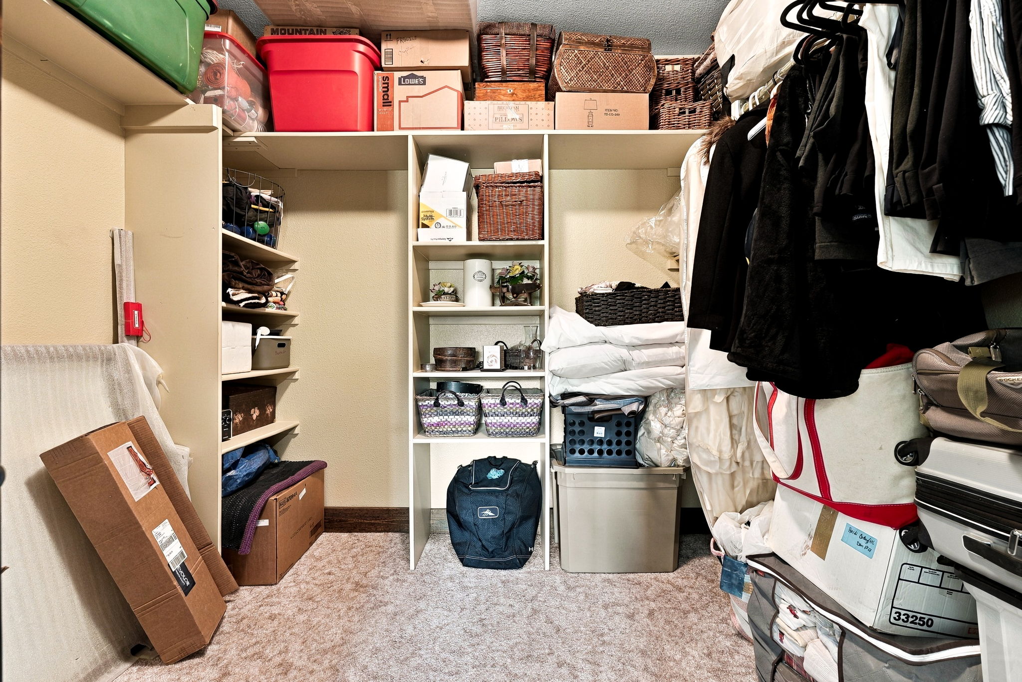 This organized walk-in closet features a combination of hanging rods, open shelving, and floor space for storage bins and boxes. The layout includes a central shelving unit flanked by hanging areas, providing ample room for clothing and personal items. The neutral walls and carpeted flooring create a functional and tidy storage environment.